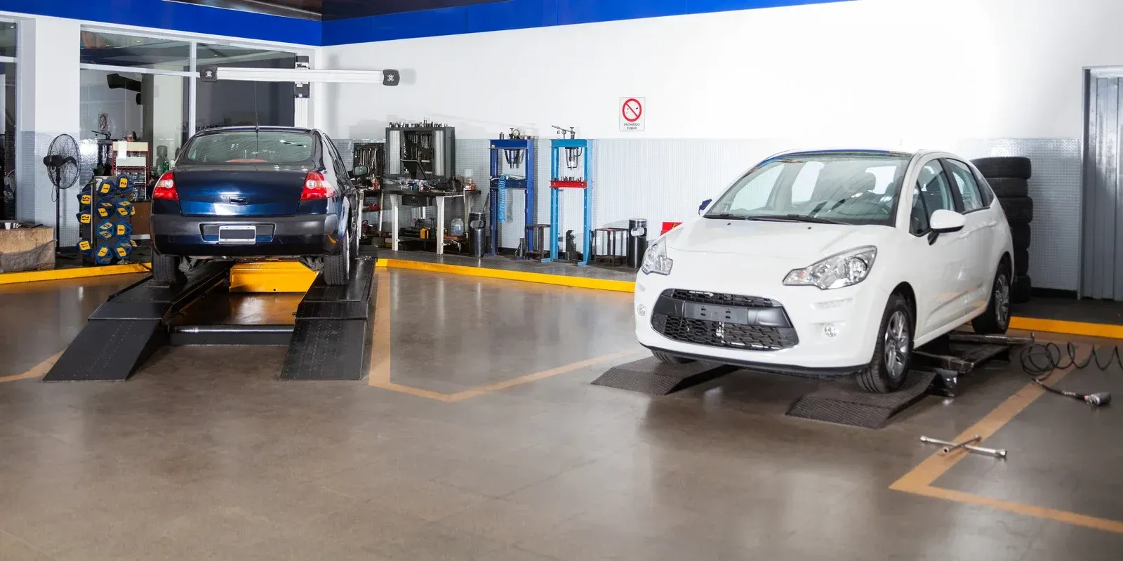 A clean, indoor auto repair shop with a dark blue car on a lift and a white car parked on a floor ramp.