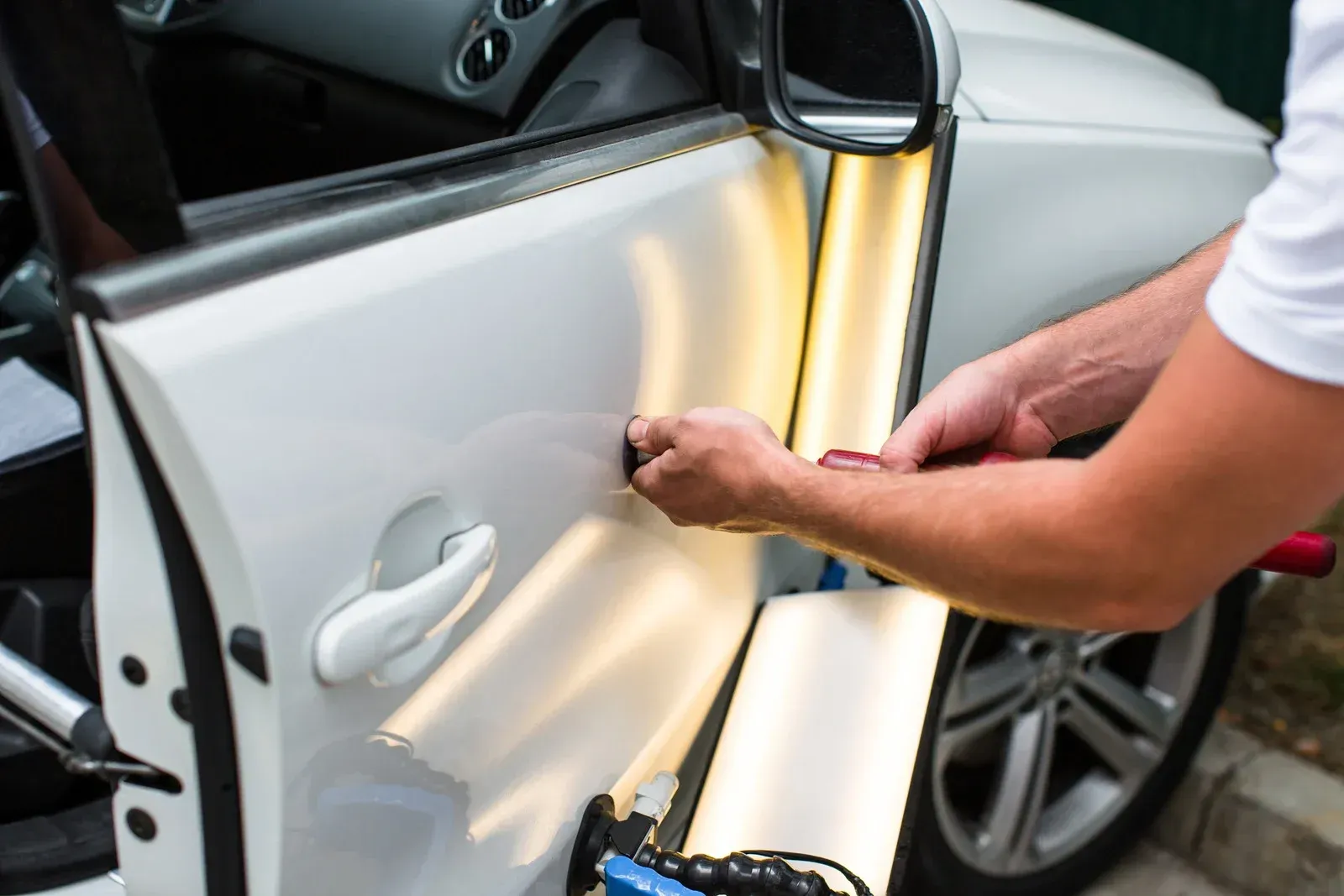 A technician using a tool to repair a dent on a white car door, illuminated by a specialized reflected light board.