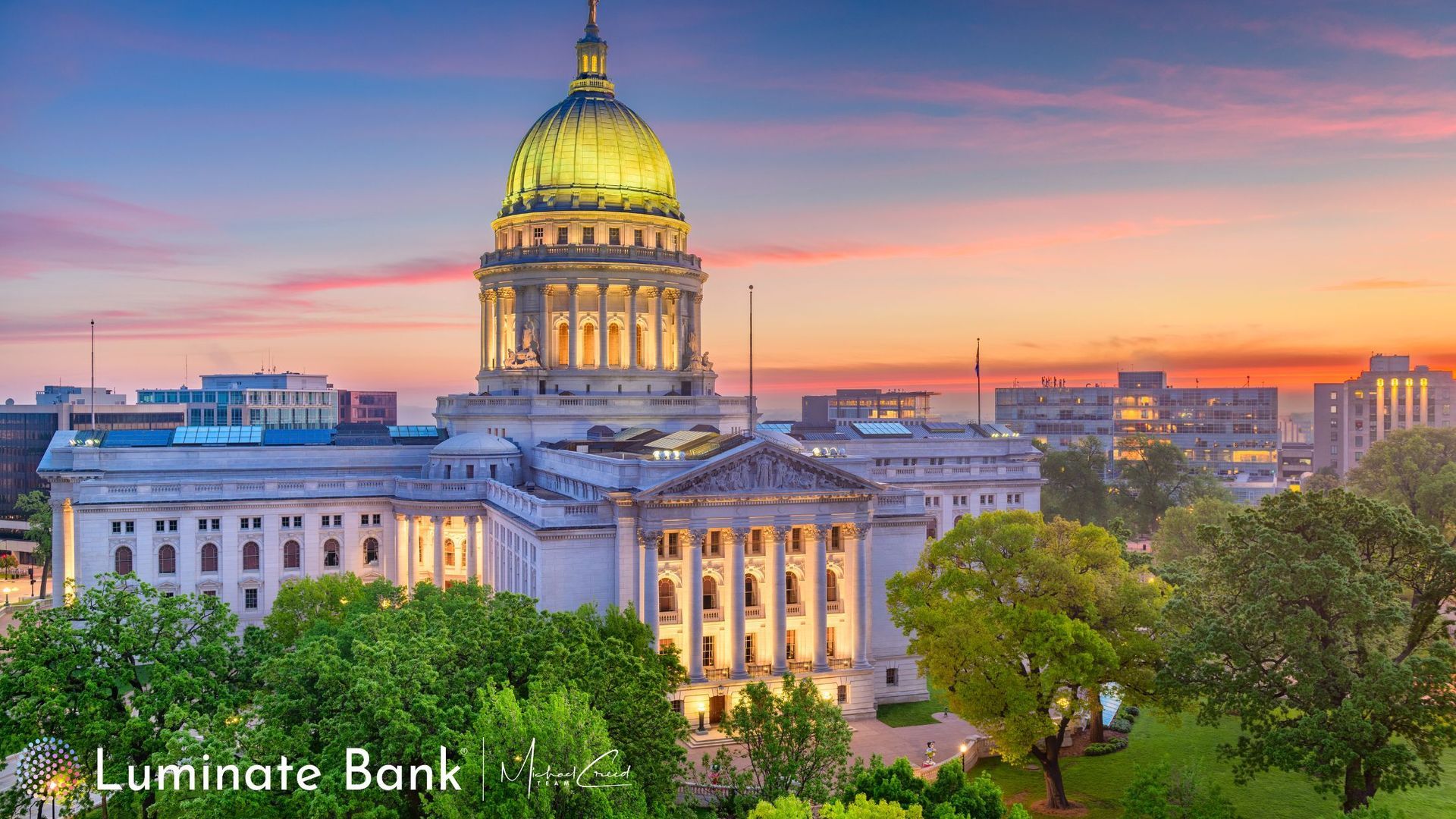 Wisconsin State Capitol building with illuminated dome at sunset. Green trees and city skyline.
