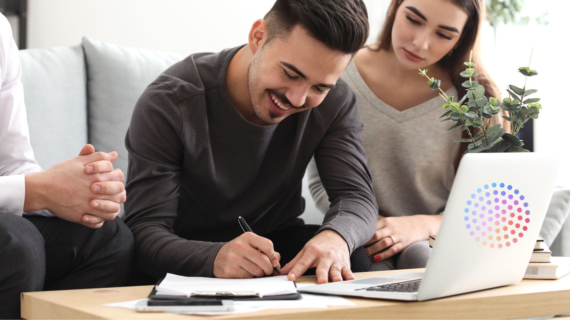 Man signing a document at a table, with a woman and another person looking on; laptop visible.