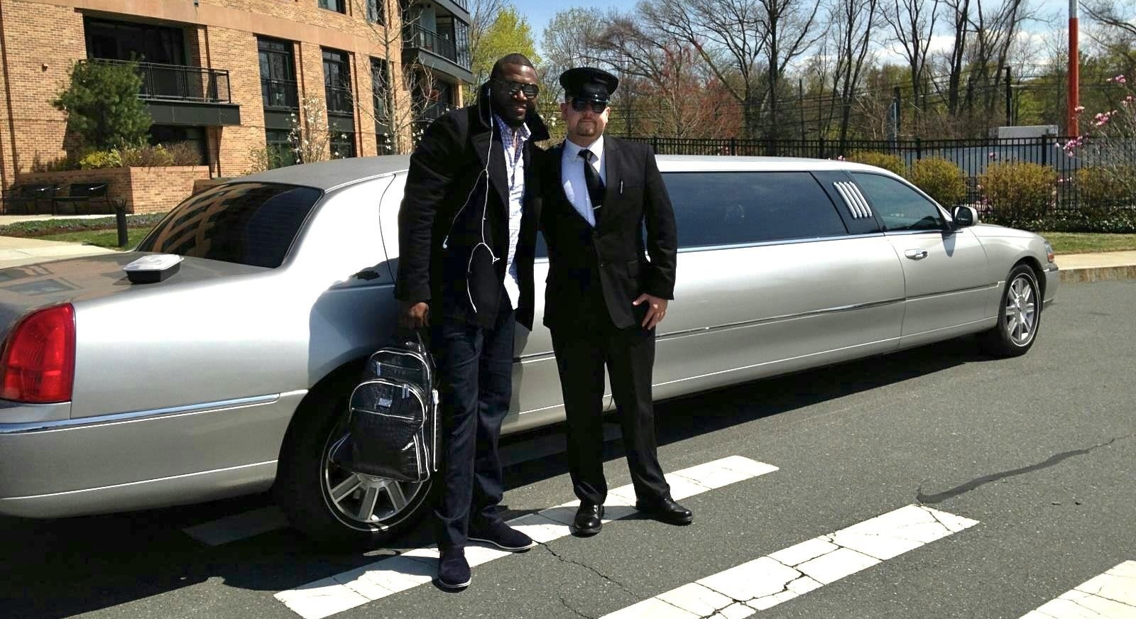 Man and chauffeur standing beside a silver limousine on a street. Brick building visible in the background.