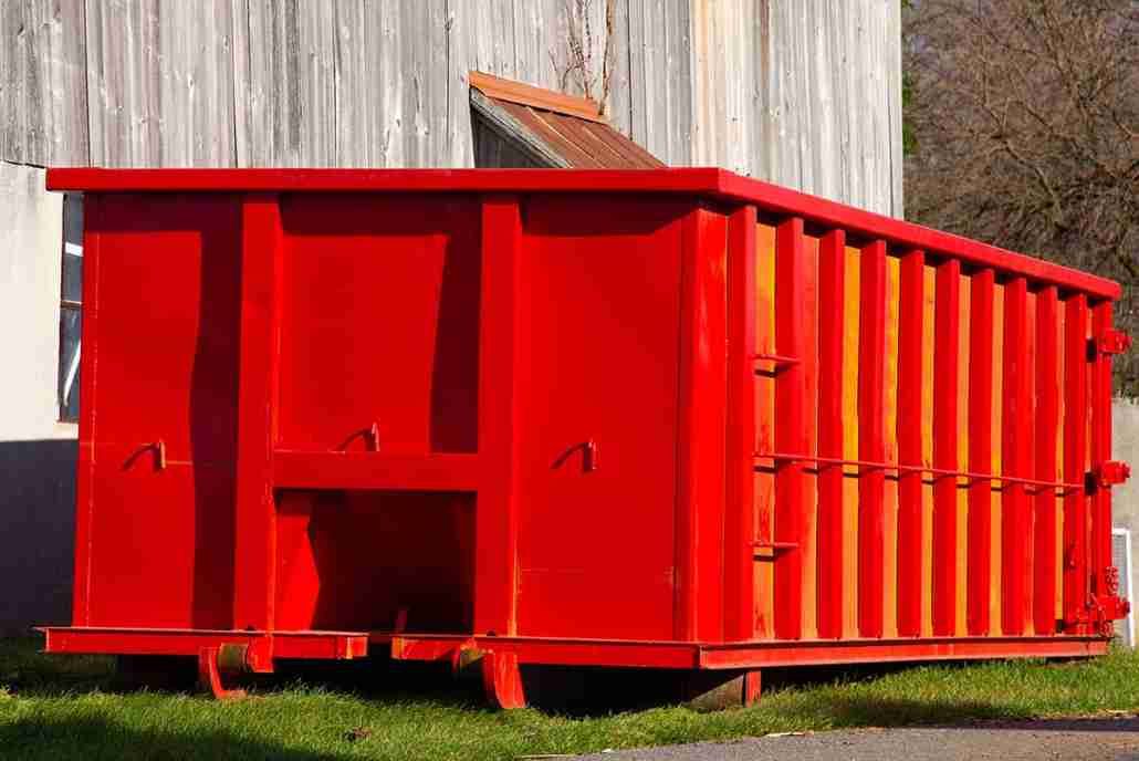 A large red dumpster is sitting in the grass in front of a building.