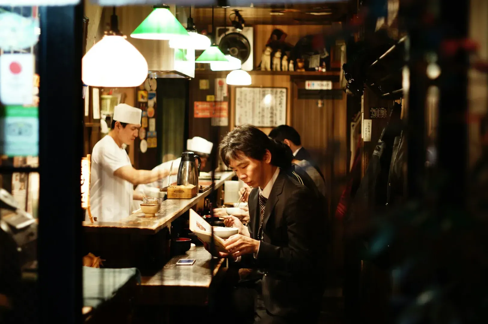 A cozy Japanese restaurant interior. A man in a suit reads a menu at the counter.