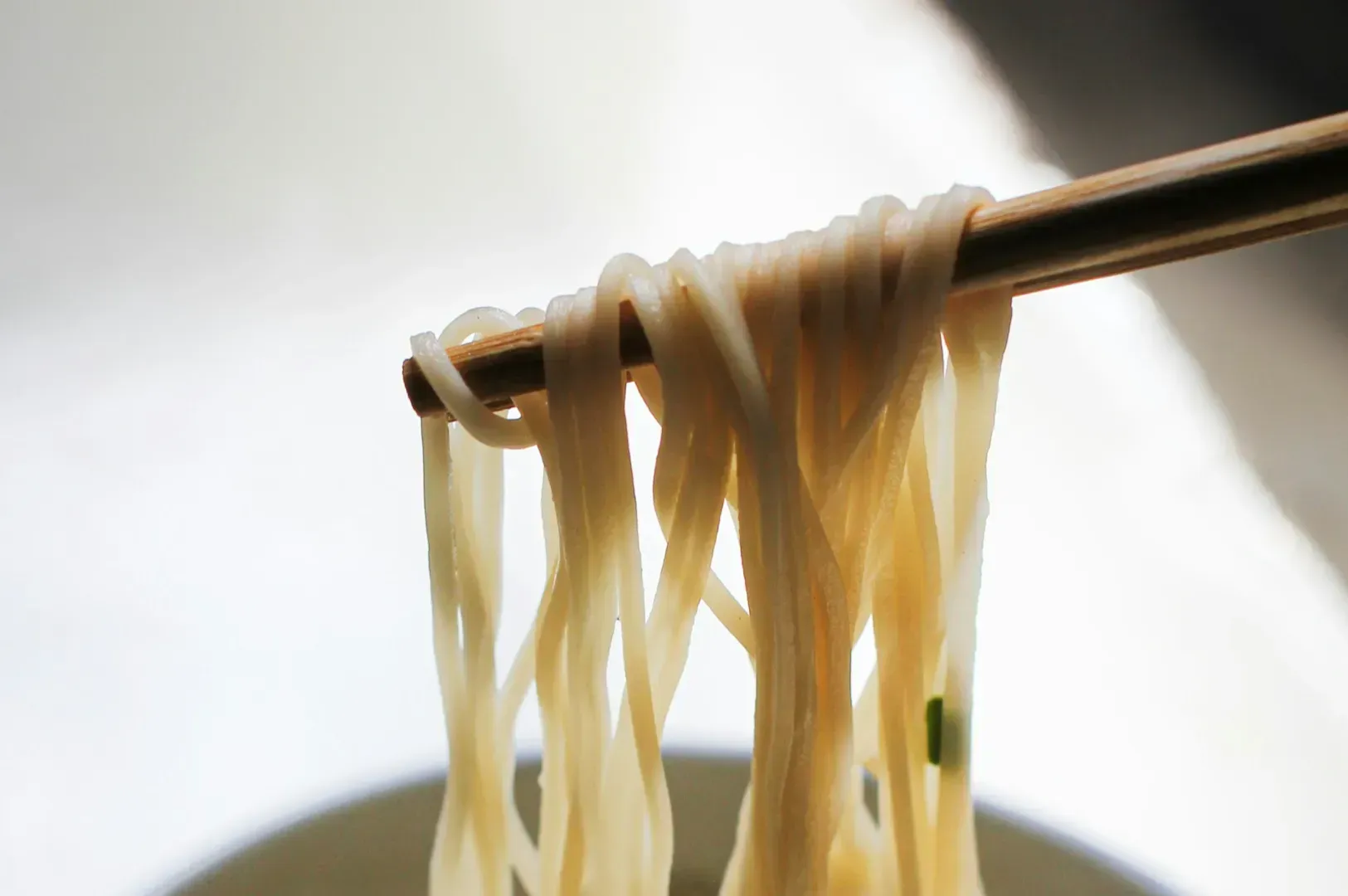 Chopsticks holding steaming, light-colored noodles above a bowl, with a soft-focus background.