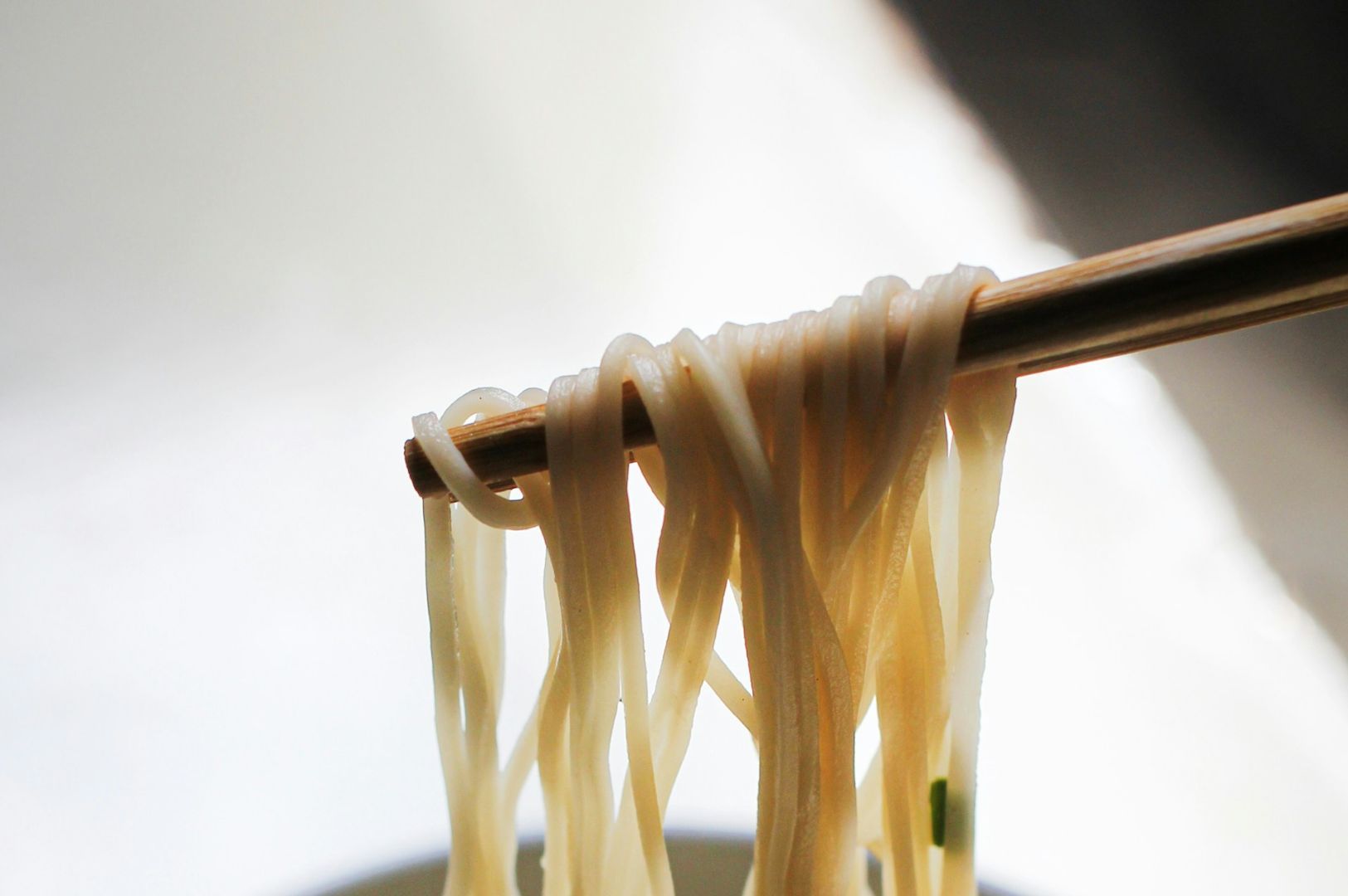 Close-up of chopsticks lifting white, glistening noodles from a bowl against a soft.