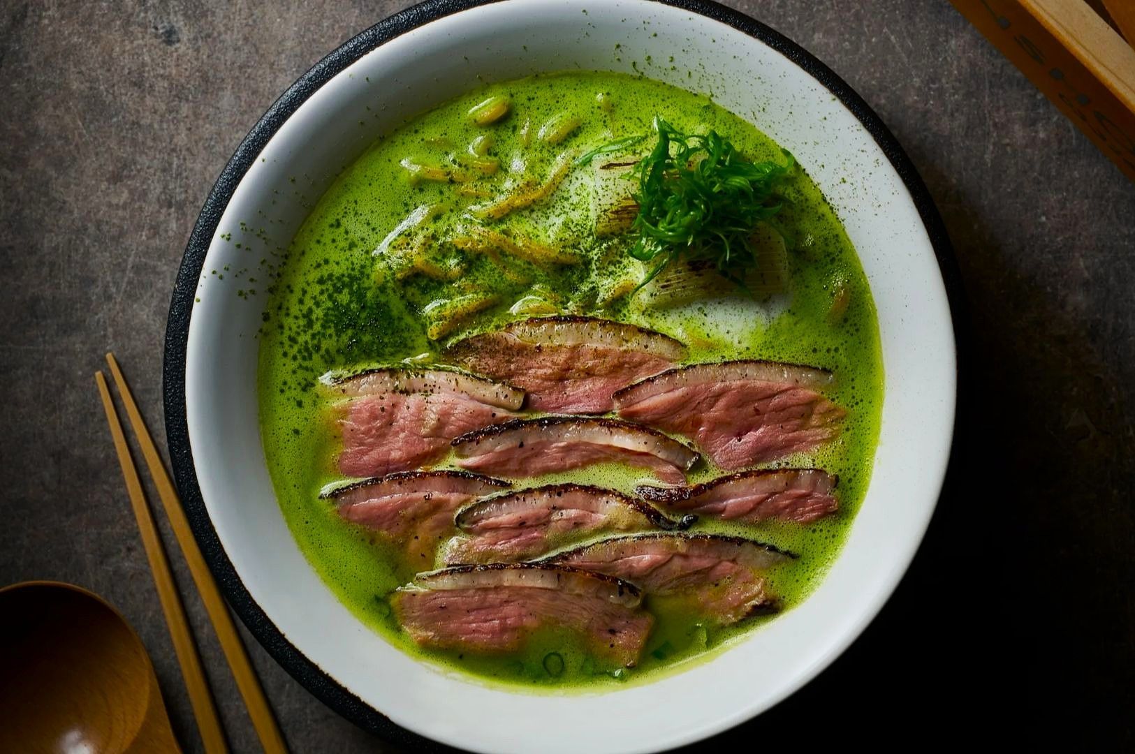 A white bowl of ramen with vibrant green broth, topped with seared slices of duck, fresh herbs,.