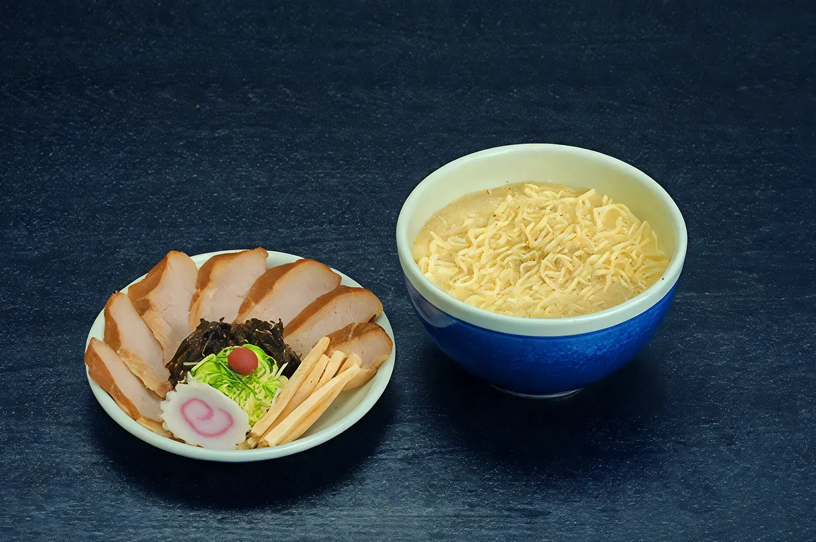 A blue bowl of plain ramen noodles next to a plate with sliced pork, seaweed, green onions.