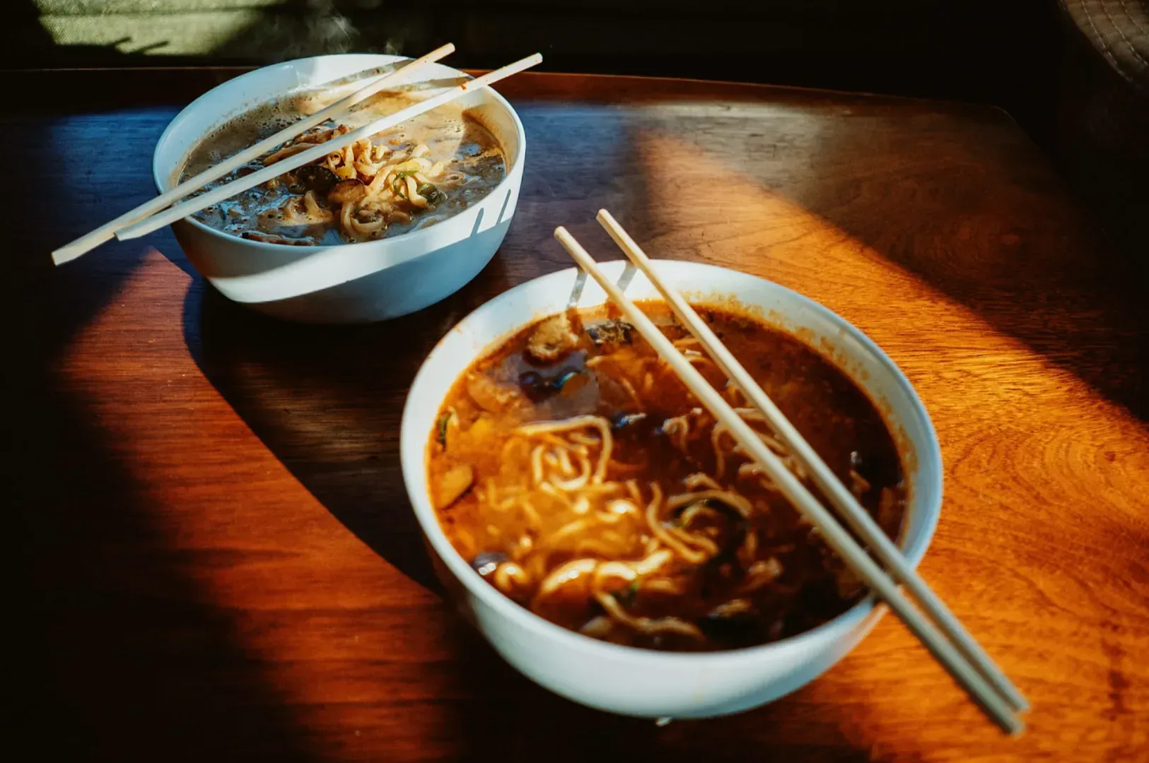 Two bowls of ramen with chopsticks rest on a wooden table. Sunlight casts a warm glow, highlighting the rich broth and noodles. Cozy and inviting scene.