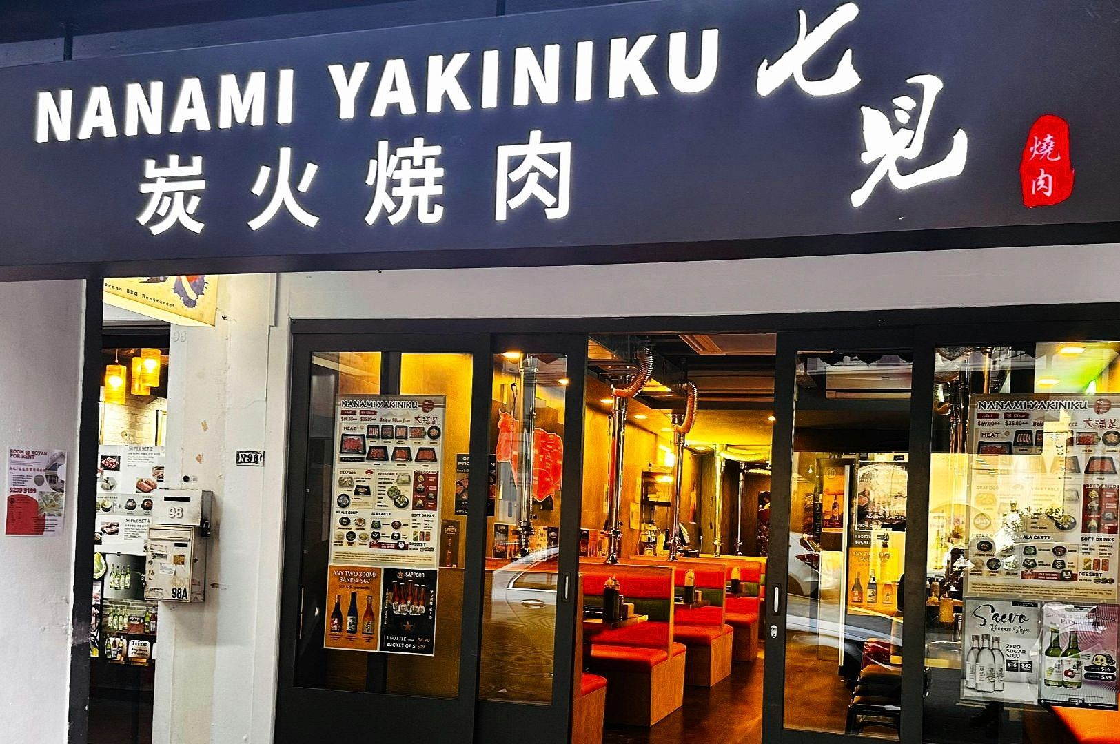 Storefront of Nanami Yakiniku, a Japanese BBQ restaurant, with a large white sign, glass entry doors, and a warmly lit interior visible through the windows.