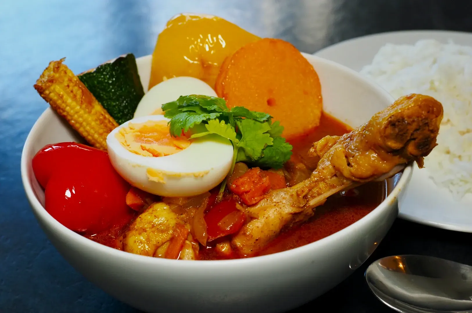 A vibrant bowl of curry featuring a chicken drumstick, boiled egg, assorted colorful vegetables.