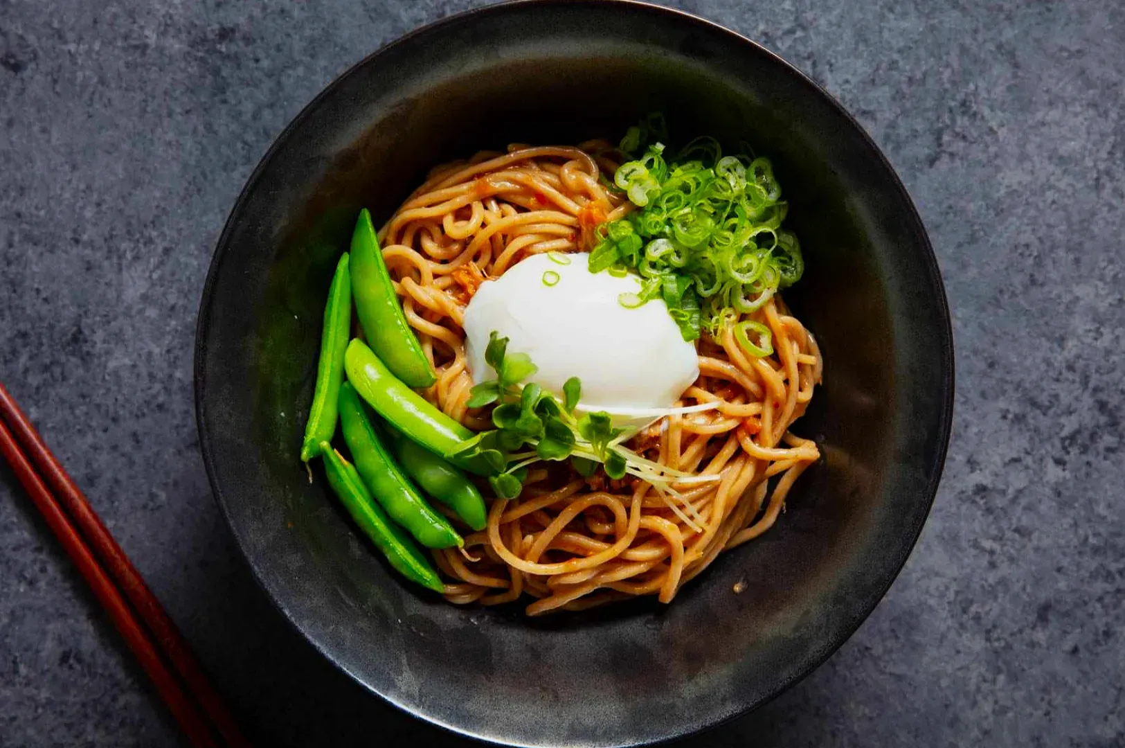 A black bowl of soba noodles topped with a poached egg, green peas, chopped scallions, and sprouts.