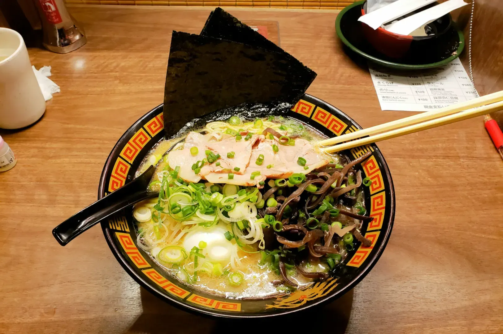 A bowl of ramen on a wooden table features sliced pork, green onions, wood ear mushrooms.