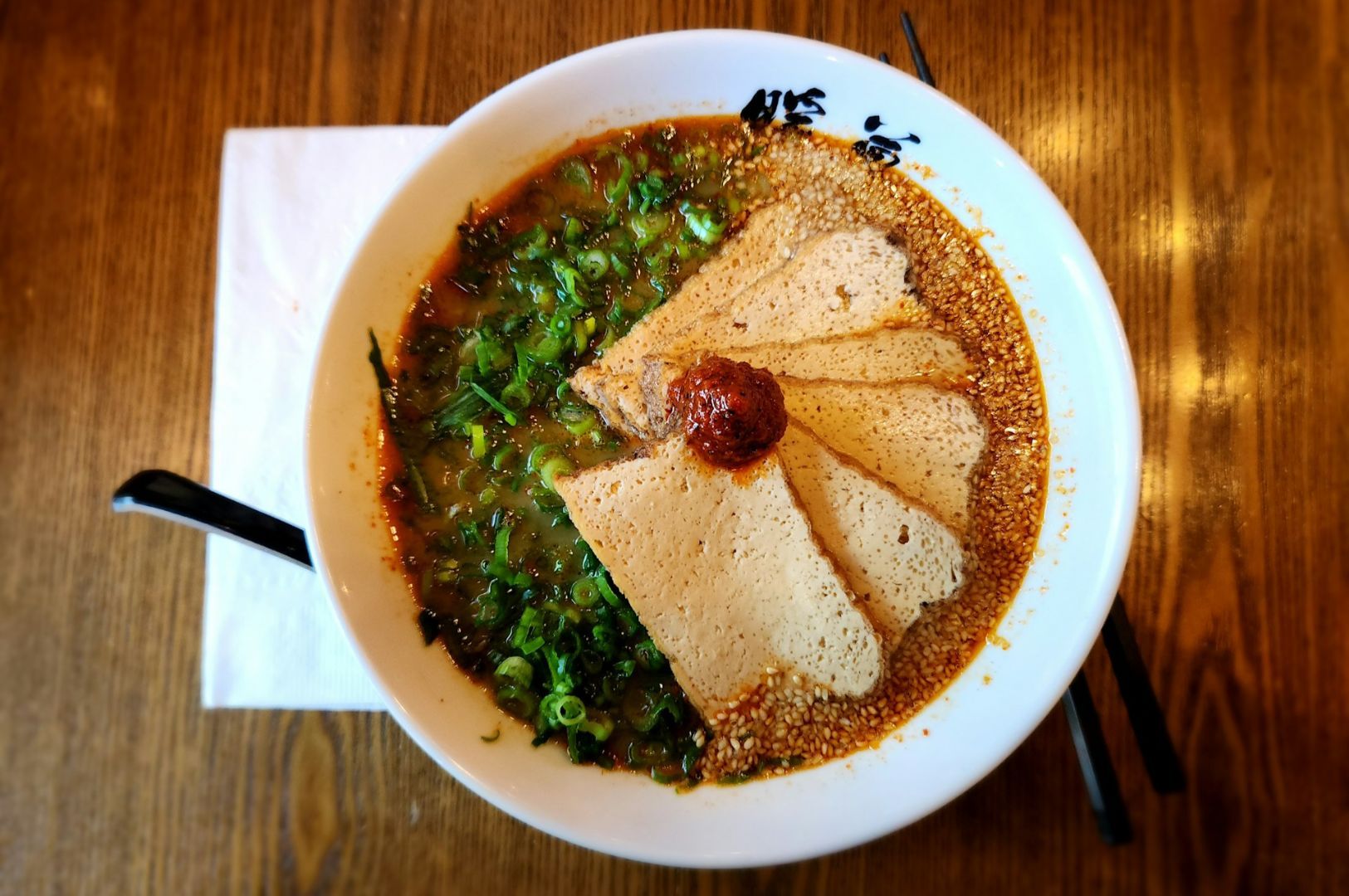 A steaming bowl of ramen on a wooden table, featuring slices of tofu, a dollop of spicy red paste.