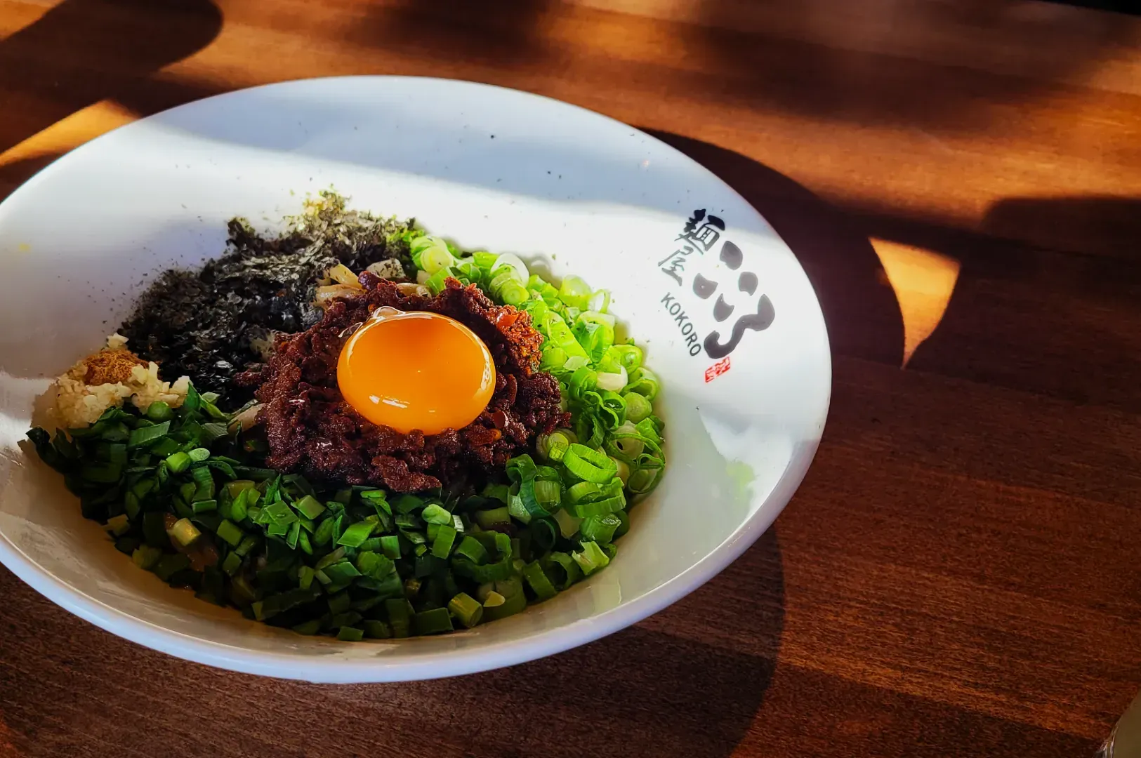 Bowl of ramen ingredients on a wooden table in sunlight. Topped with raw egg yolk, minced meat.
