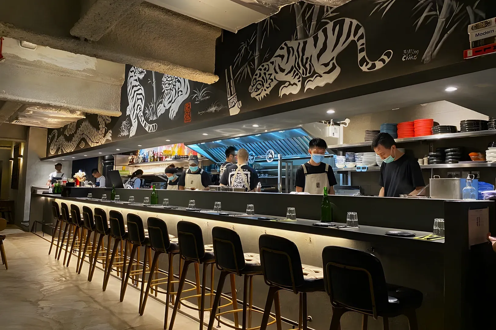 A modern restaurant interior features a long bar with high stools. Chefs work in an open kitchen.