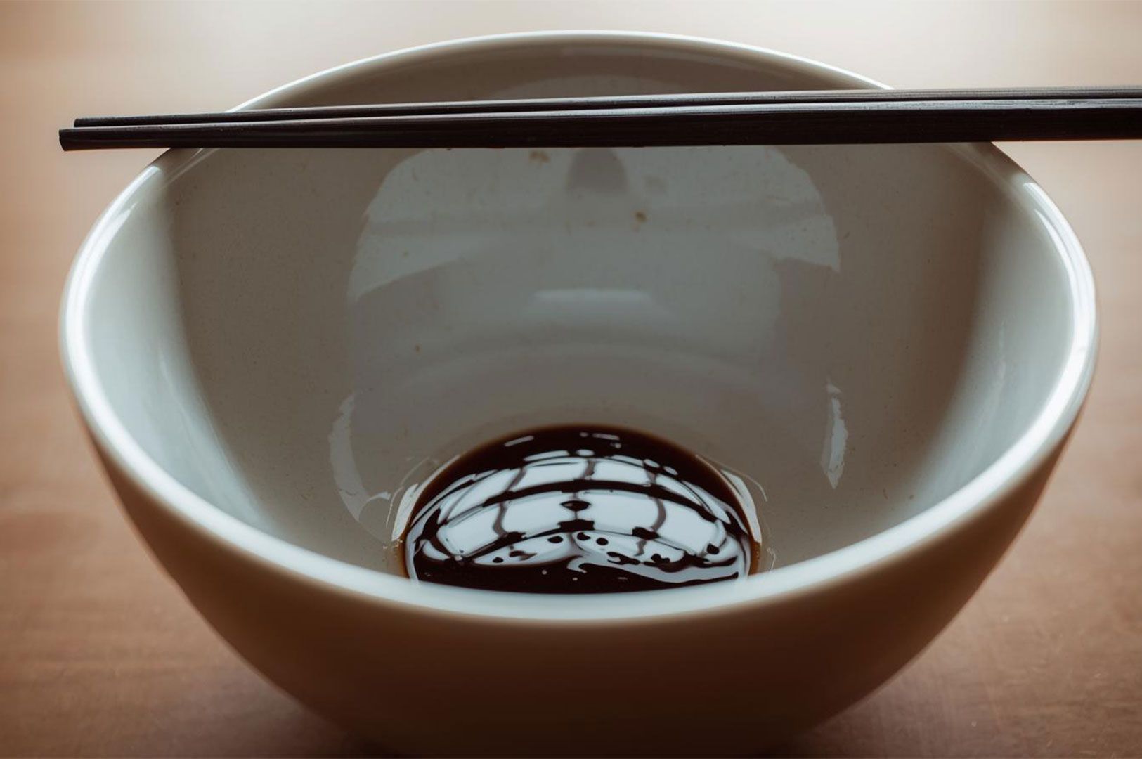 A small pool of dark, glossy Shoyu (soy sauce) tare reflecting the chopsticks and bowl rim,.