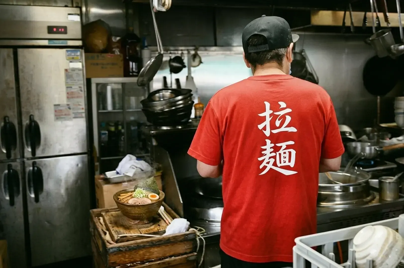 Back view of a ramen chef in a red shirt working in a kitchen with a prepared bowl ready for service