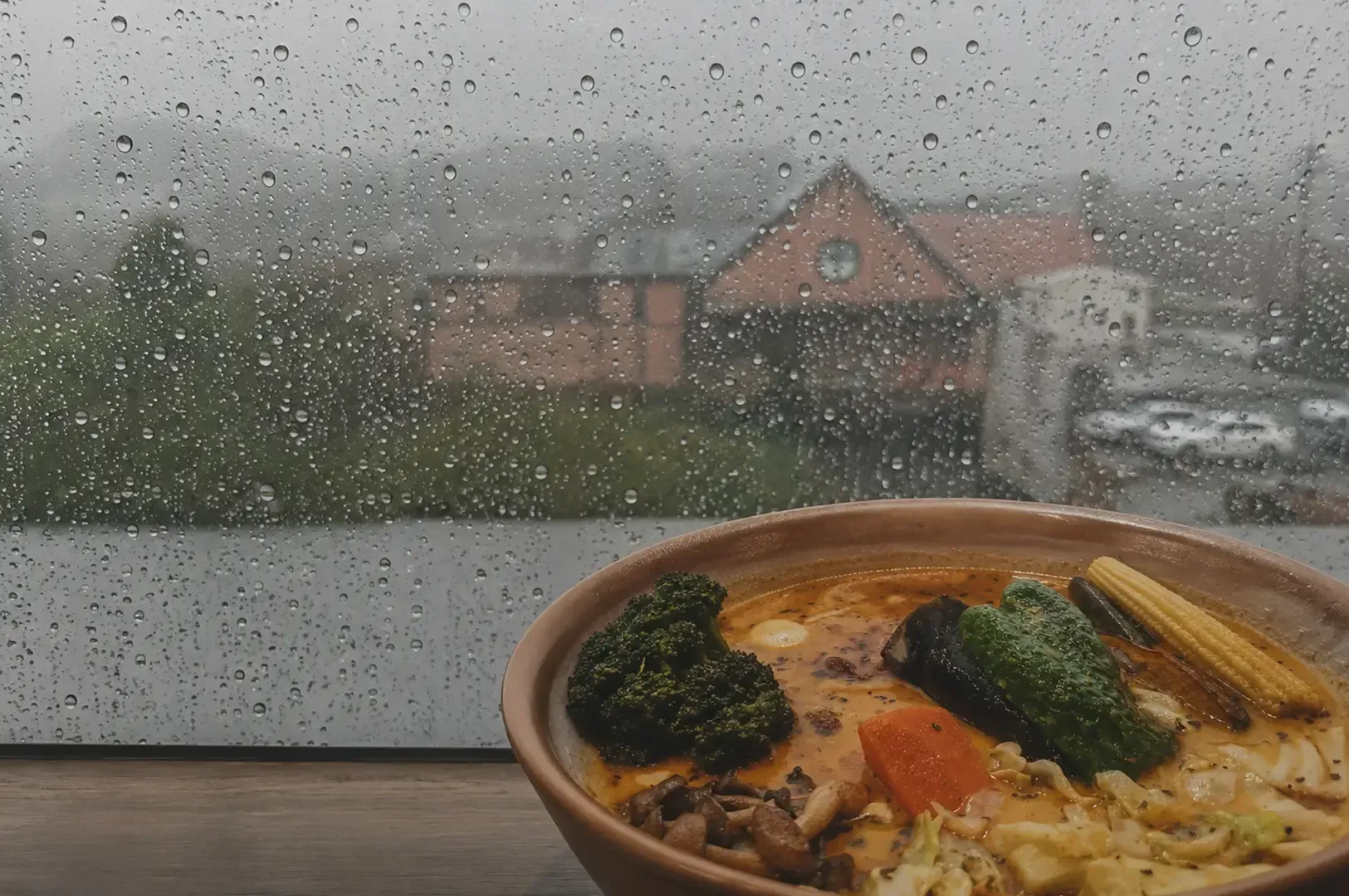 Bowl of vegetable soup curry on a table in front of a rain-streaked window with a blurred rainy view