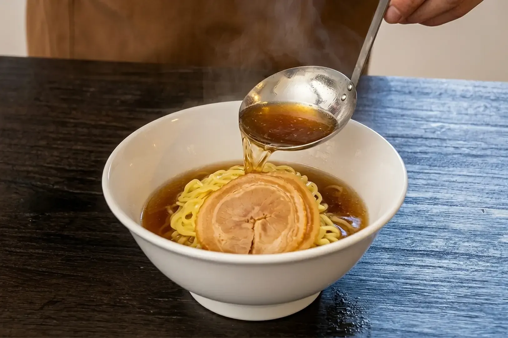 Ladling clear shoyu broth over curly noodles and chashu pork in traditional ramen bowl