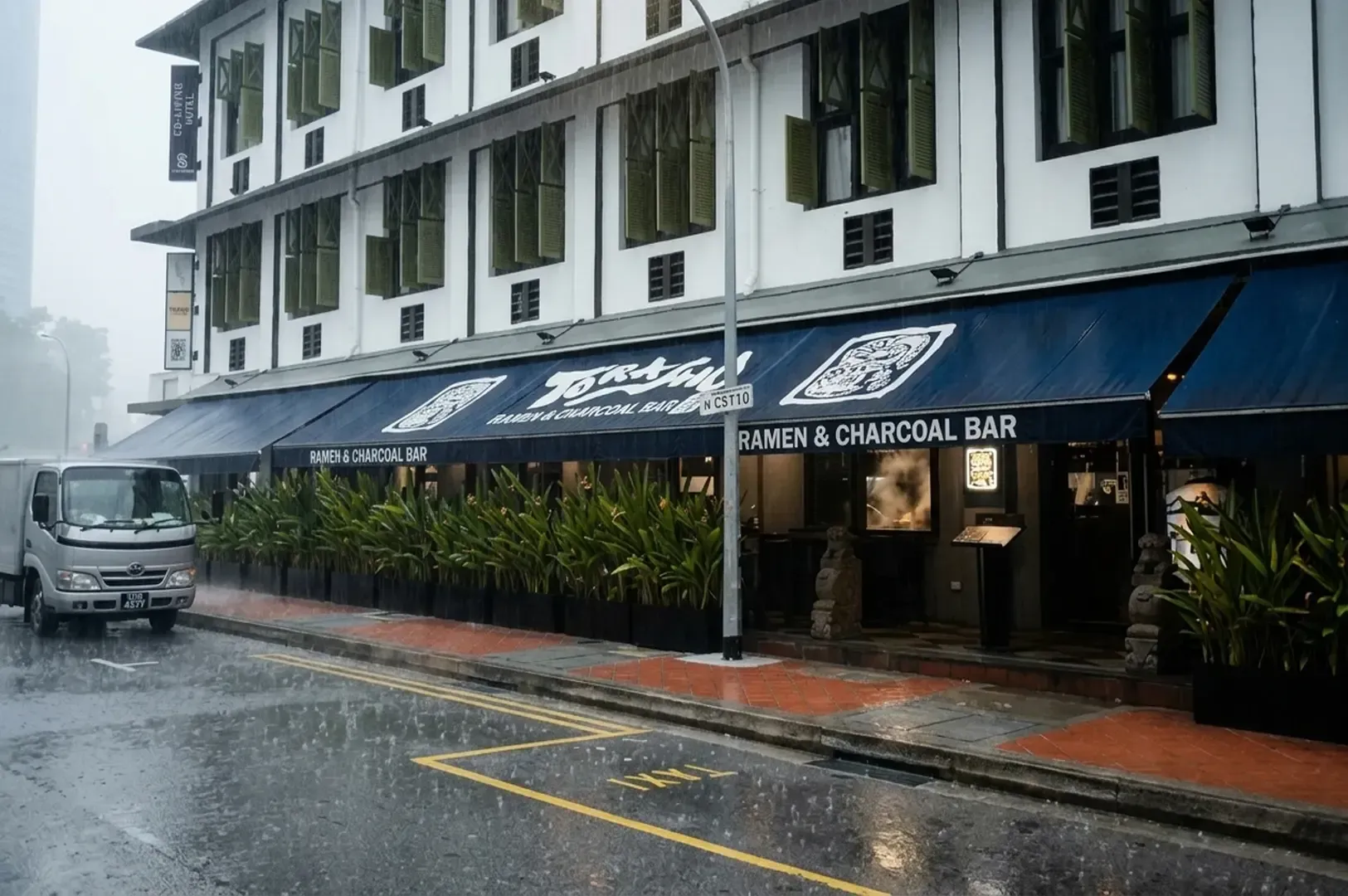 Street view of Torasho Ramen & Charcoal Bar with blue awnings and green shutters on a rainy day