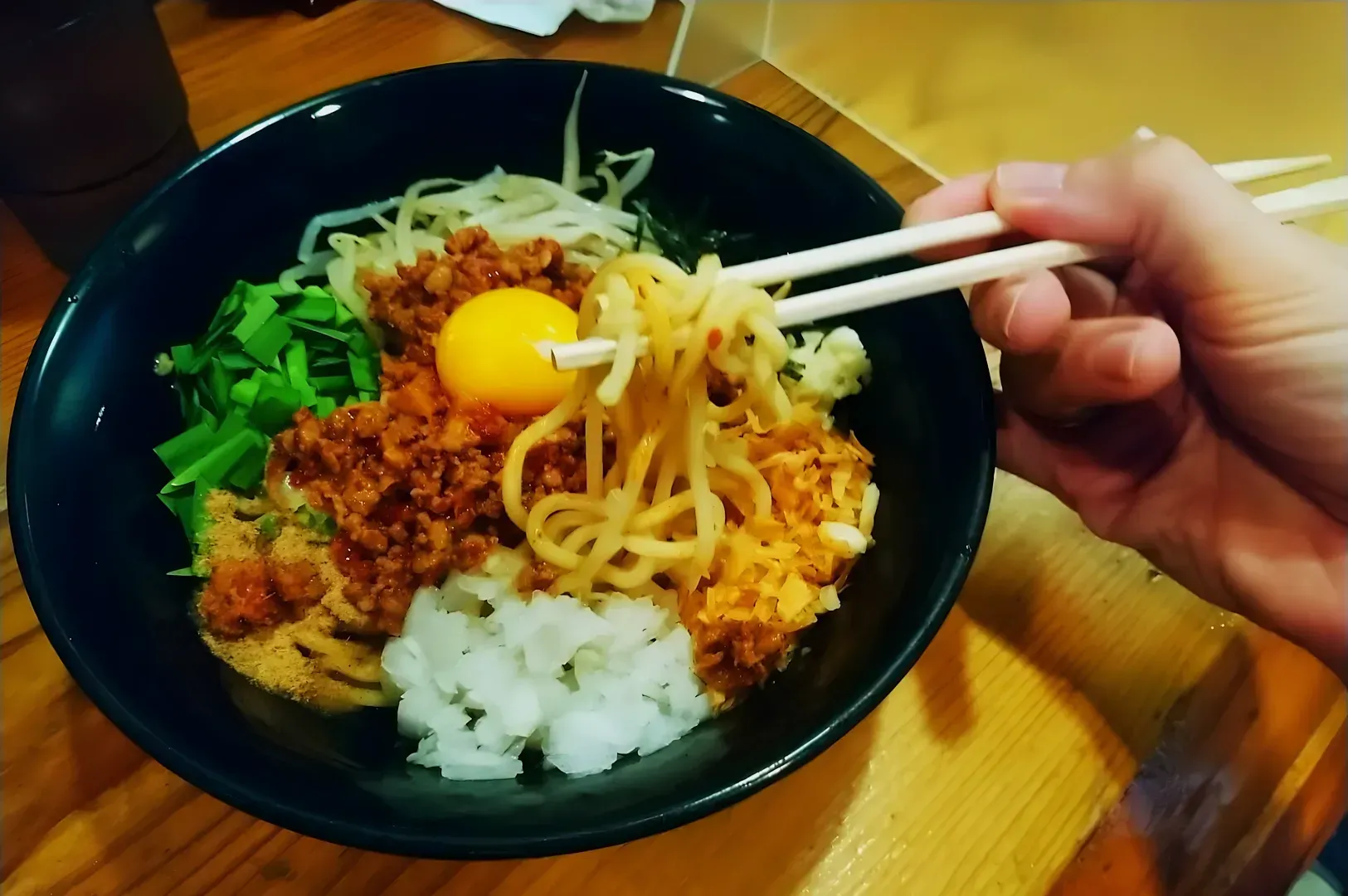 A hand using chopsticks to lift noodles from a black bowl filled with colorful ingredients including a raw egg yolk, minced meat, and vegetables.