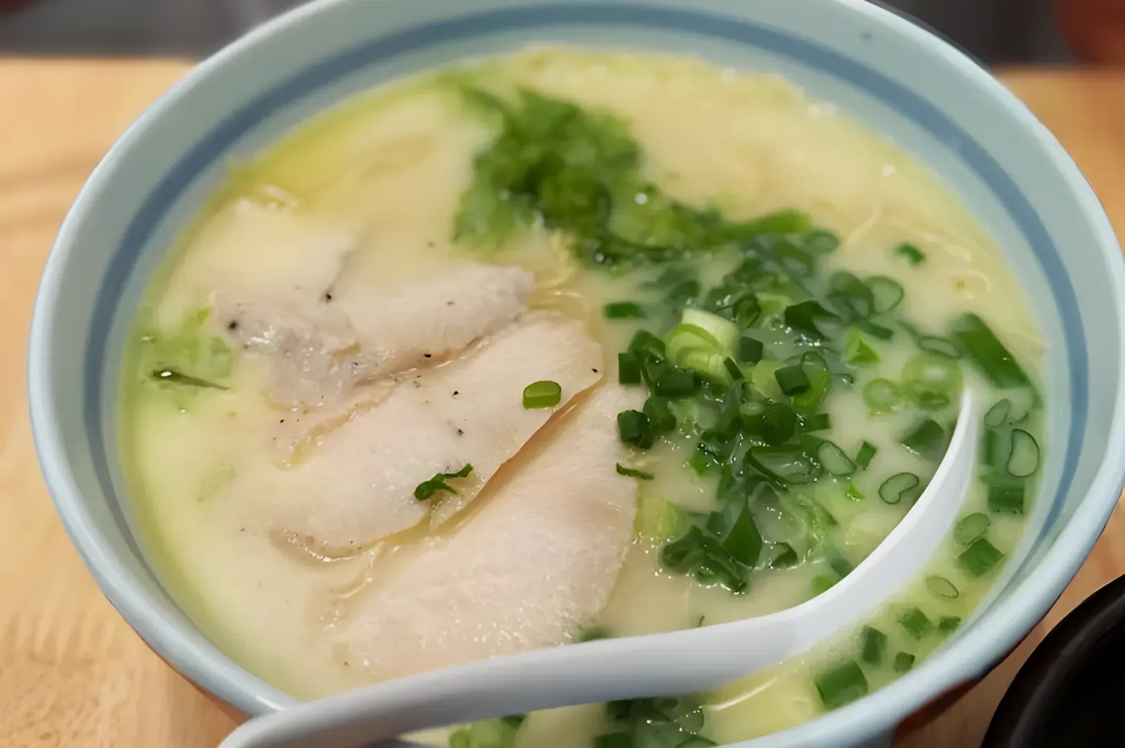 A steaming bowl of creamy ramen soup with slices of tender chicken, chopped green onions, and a white spoon. The dish appears savory and comforting.