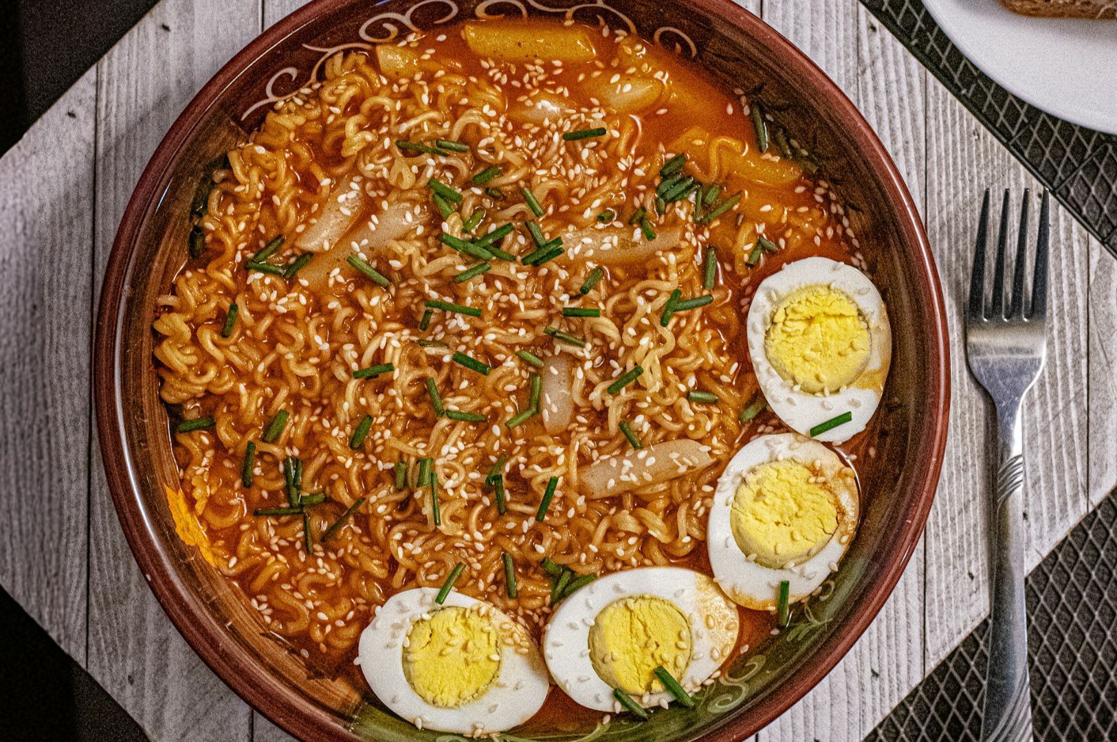 A bowl of spicy ramen topped with sliced boiled eggs, chopped green onions, and sesame seeds, on a wooden table next to a fork. The scene appears appetizing and rich in flavor.