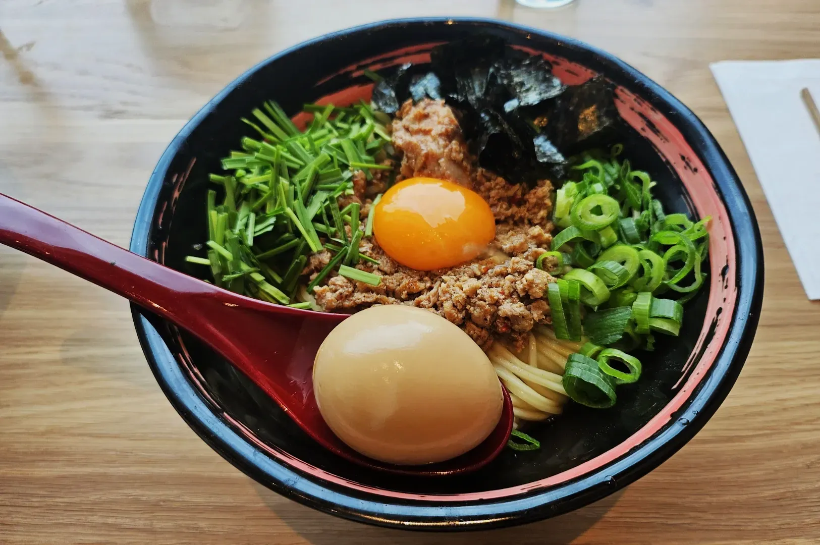 A bowl of ramen with green onions, chives, ground meat, a raw egg yolk, seaweed, and a boiled egg in a red spoon, on a wooden table.