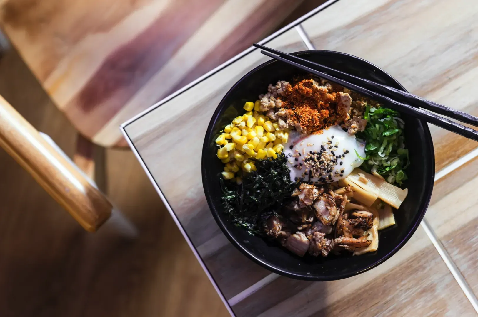 A bowl of ramen with an egg, corn, seaweed, bamboo shoots, and spices is on a wooden table, with chopsticks resting on the bowl. Warm, inviting atmosphere.