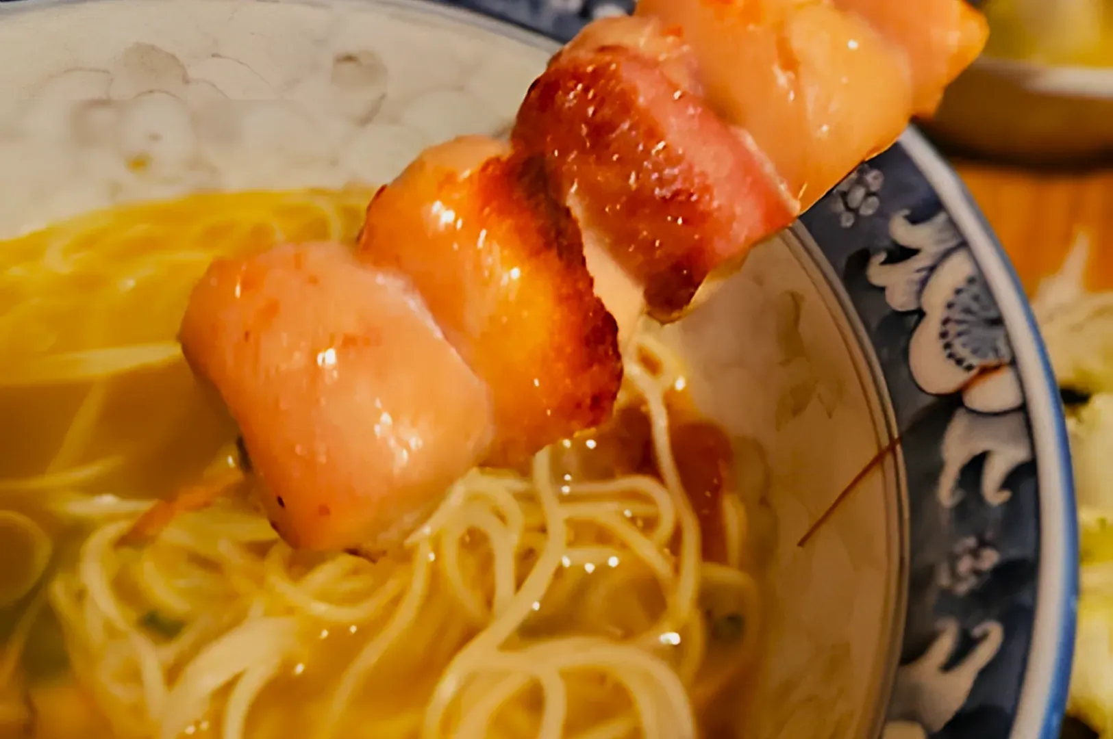 Close-up of a skewer with grilled chicken placed over a bowl of ramen in light broth, set in a decorative blue and white bowl, suggesting warmth and comfort.