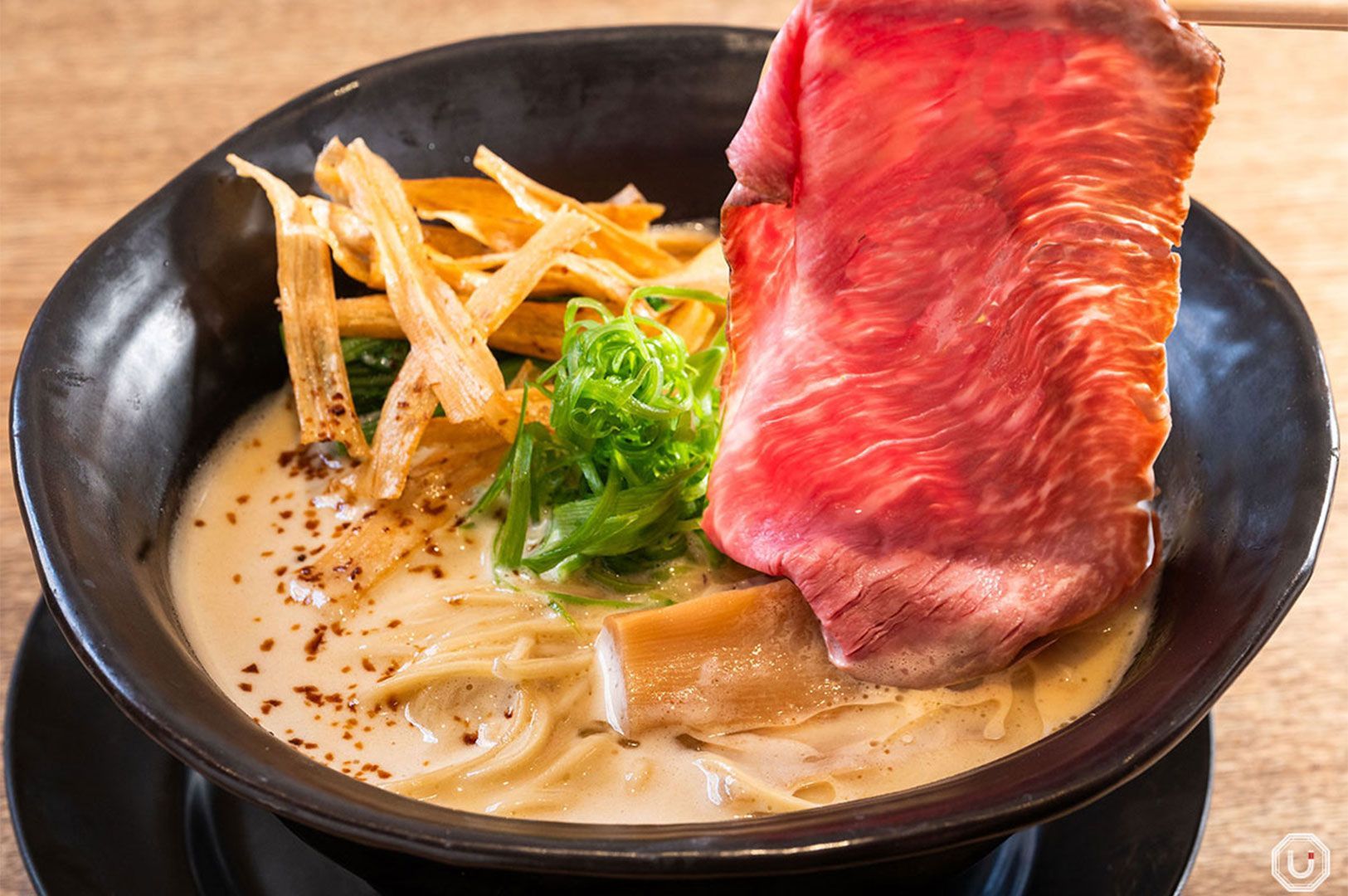 Close-up of a hand lifting a thin slice of raw-seared wagyu beef out of a bowl of creamy white ramen broth with noodles and bamboo shoots.