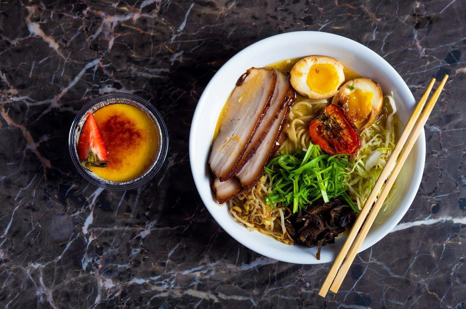 A bowl of ramen with sliced pork, soft-boiled eggs, green onions, and tomato, next to a crème brûlée dessert topped with a strawberry, on a marble table.