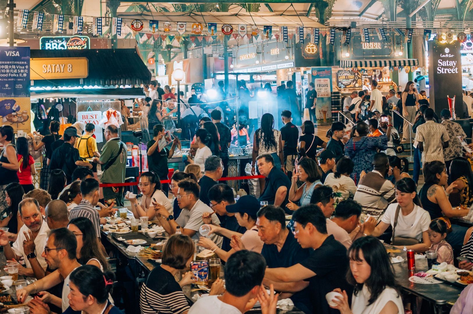 A bustling food market scene with diverse groups of people eating at crowded communal tables under vibrant signage. The atmosphere is lively and festive.