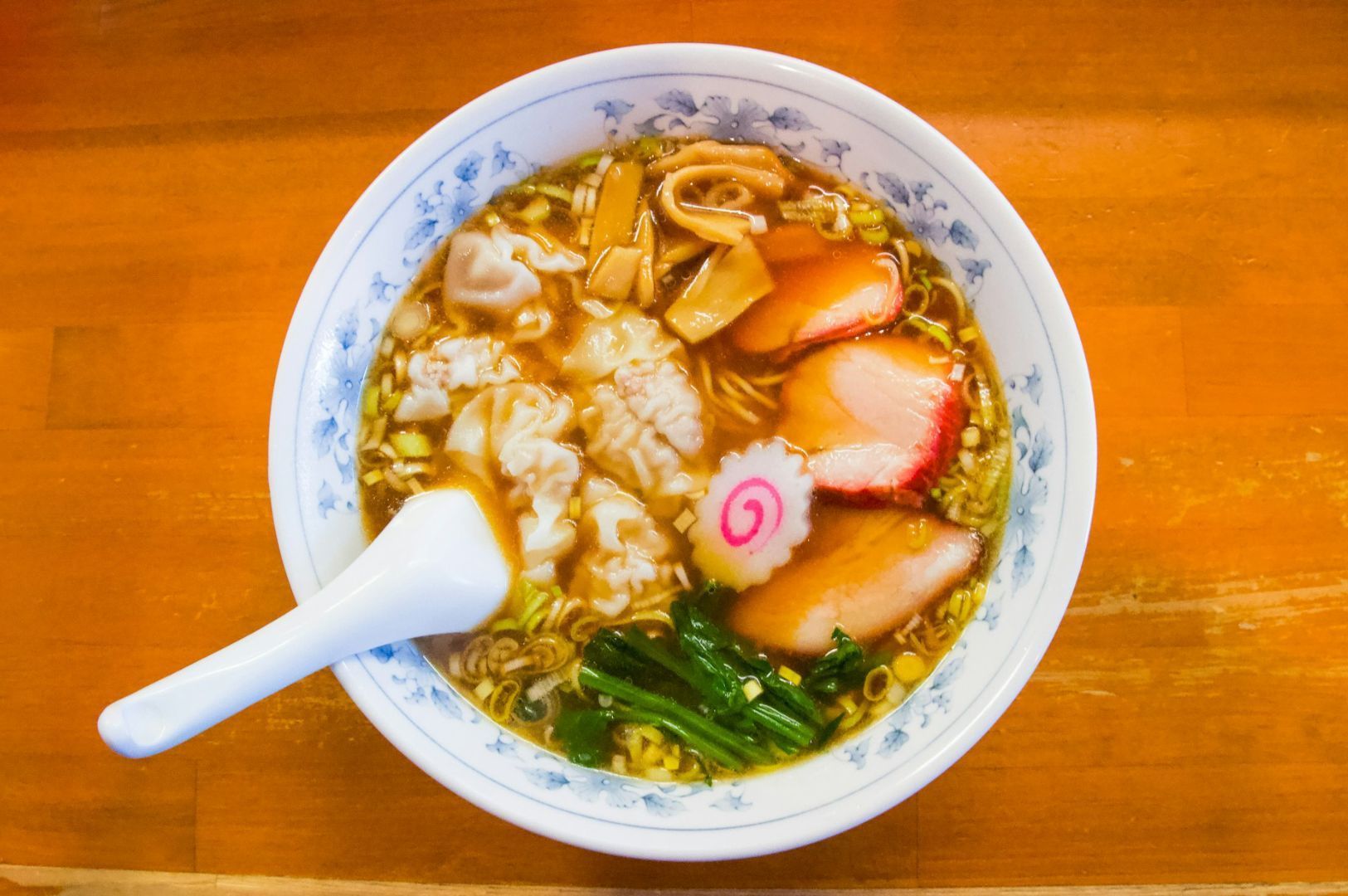 A steaming bowl of ramen on a wooden table with slices of pork, bamboo shoots, spinach, dumplings, and a narutomaki swirl, accompanied by a white spoon.