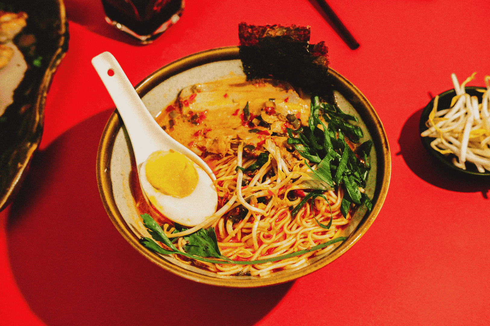 A vibrant bowl of Singapore-style ramen served on a red tabletop, featuring yellow noodles in creamy broth swirled with red chili oil, topped with a halved soft-boiled egg, leafy greens, seaweed, and a white ceramic spoon. Beside it, a small dish of bean sprouts and a glass of dark beverage add depth to the dining scene, with black chopsticks in the background. The composition highlights bold flavors and visual contrast, capturing the sensory richness of Singapore’s ramen culture.