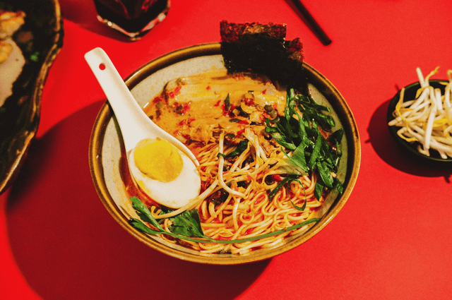 A vibrant bowl of Singapore-style ramen served on a red tabletop, featuring yellow noodles in creamy broth swirled with red chili oil, topped with a halved soft-boiled egg, leafy greens, seaweed, and a white ceramic spoon. Beside it, a small dish of bean sprouts and a glass of dark beverage add depth to the dining scene, with black chopsticks in the background. The composition highlights bold flavors and visual contrast, capturing the sensory richness of Singapore’s ramen culture.