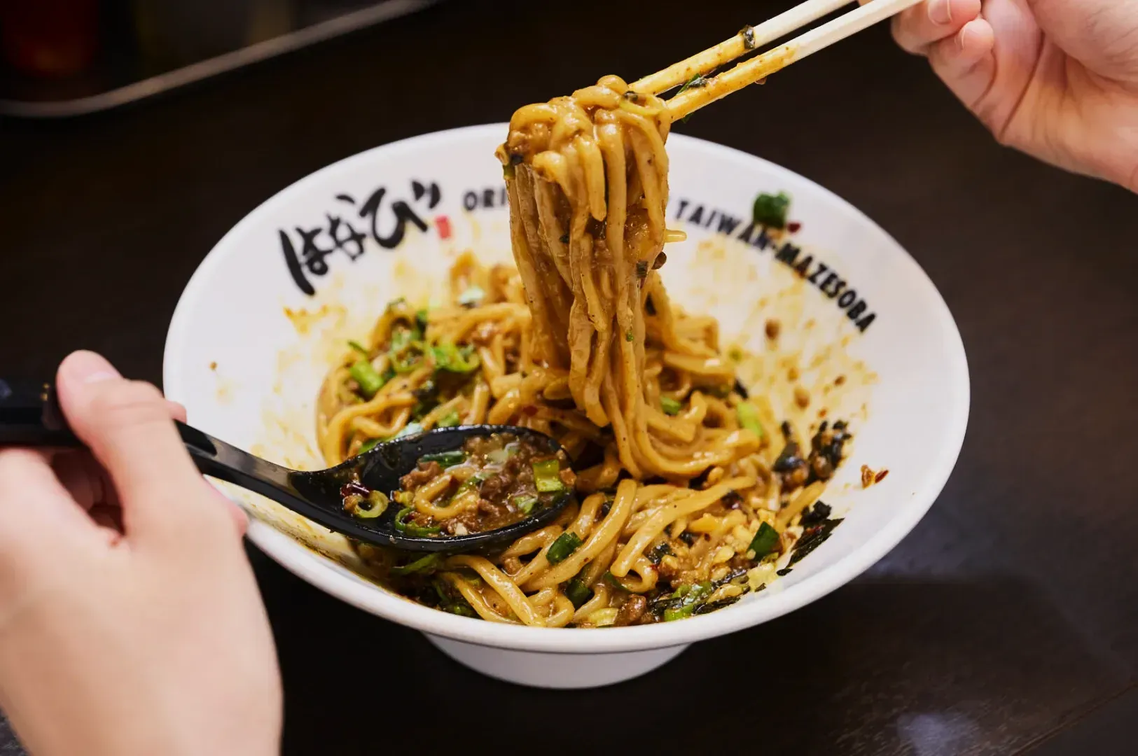 A hand holds chopsticks lifting saucy, thick udon noodles from a bowl, with another hand using a spoon. The dish includes green onions and bits of meat.