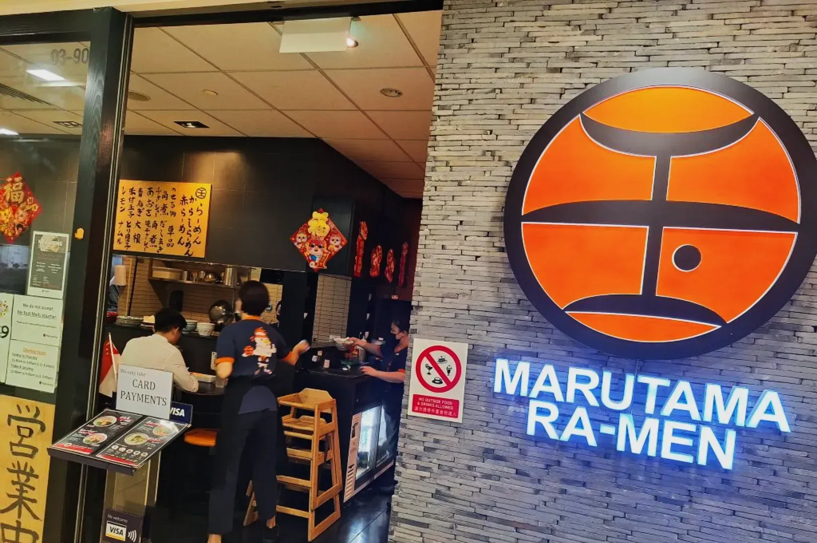 Front of a Marutama Ra-Men restaurant, featuring a bright orange logo and sign. Inside, diners enjoy ramen at a cozy counter. Warm, inviting atmosphere.