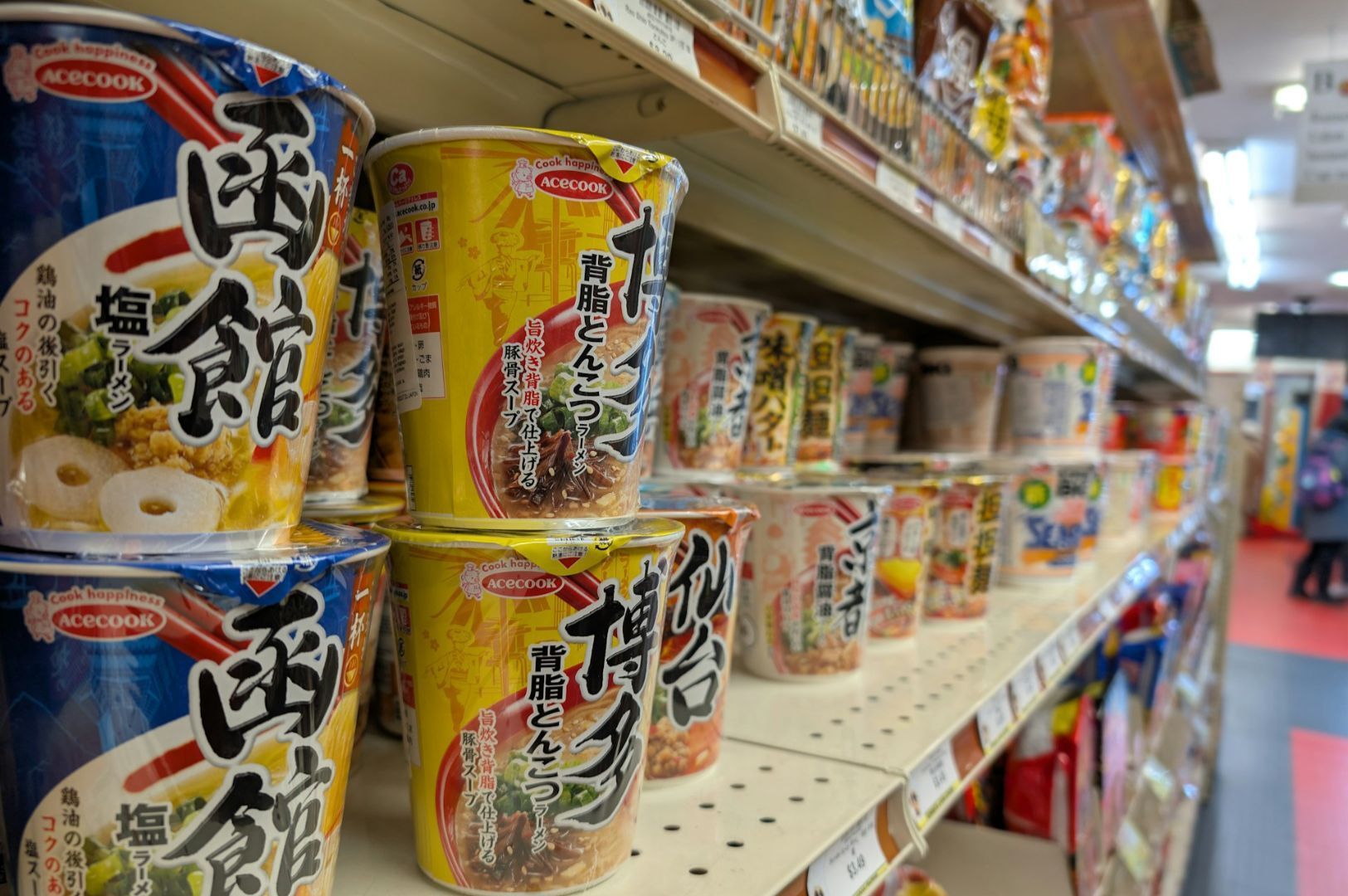 A grocery aisle filled with colorful instant noodle cups, predominantly in blue and yellow packaging with Japanese text, under bright lighting.