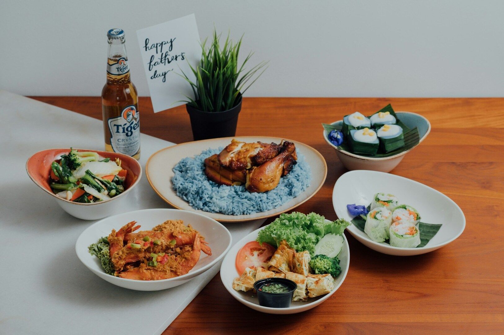 A variety of Asian dishes on a wooden table, including blue rice with grilled chicken, sushi, spring rolls, vegetables, shrimp, and a beer bottle. A 