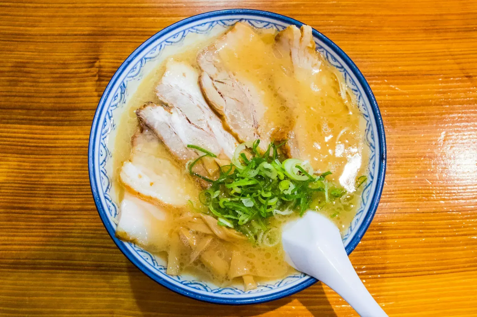 A bowl of ramen with rich broth, topped with sliced pork, bamboo shoots, and green onions. A white spoon rests in the bowl, on a wooden table.