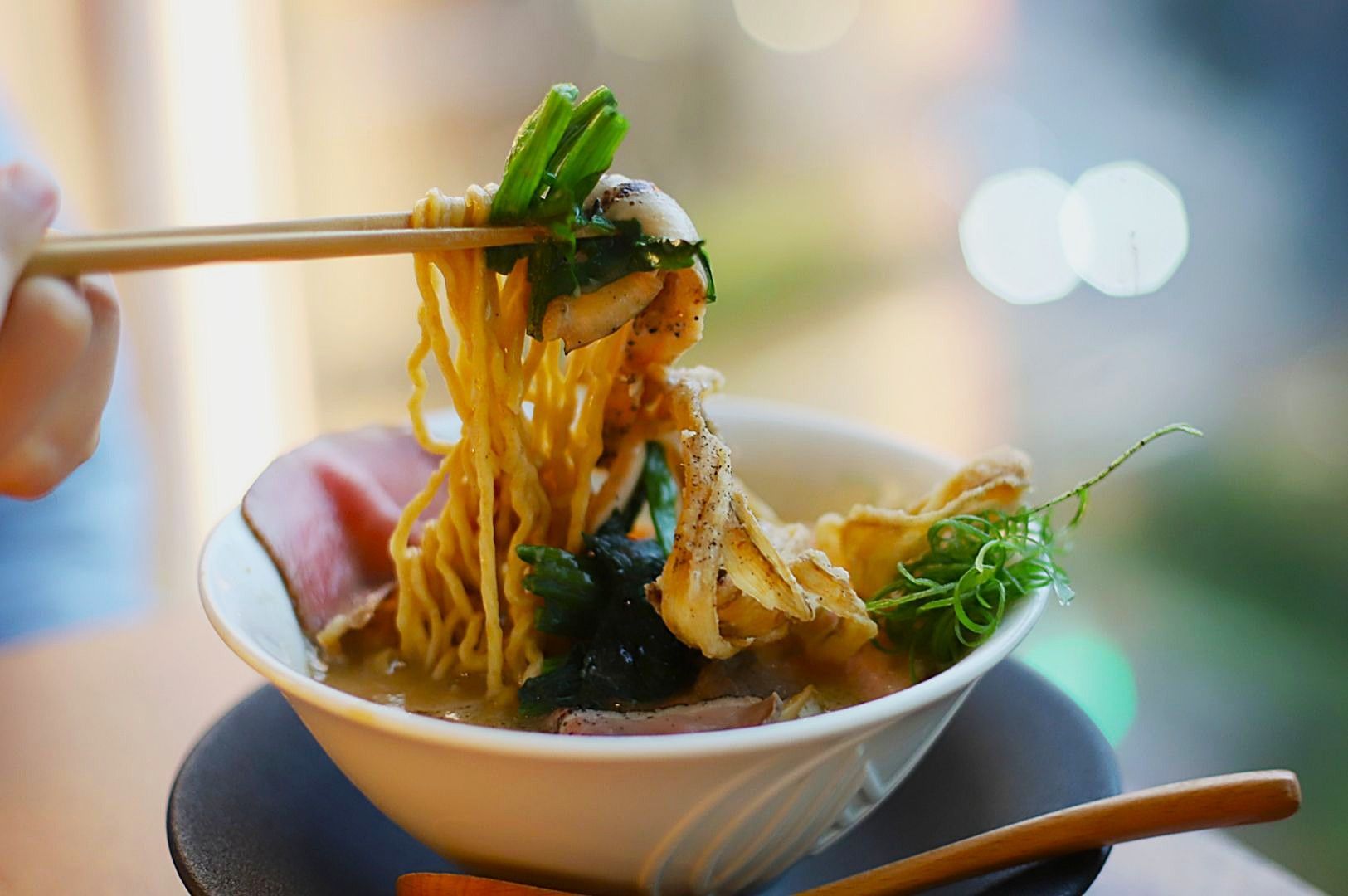 A steaming bowl of ramen is topped with grilled chicken, green vegetables, and mushrooms. Chopsticks lift noodles, set against a blurred cityscape background.