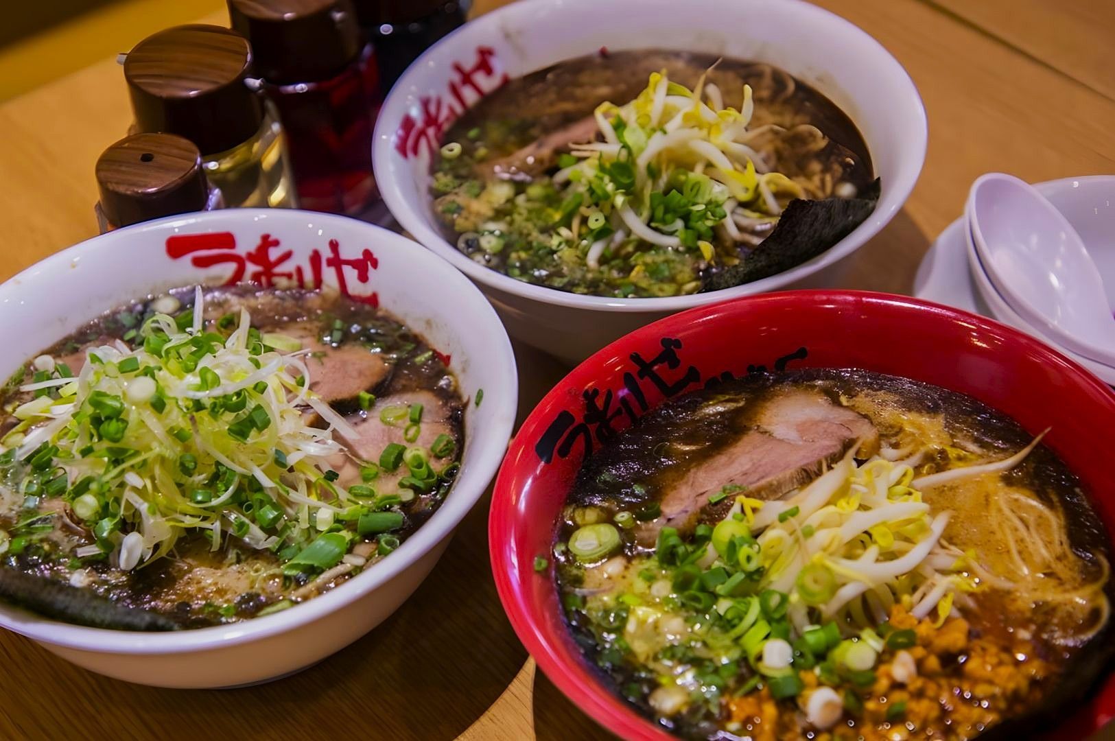 Three bowls of ramen sit on a wooden table, each topped with sliced pork, bean sprouts, and green onions. Bottles of seasoning are placed nearby.
