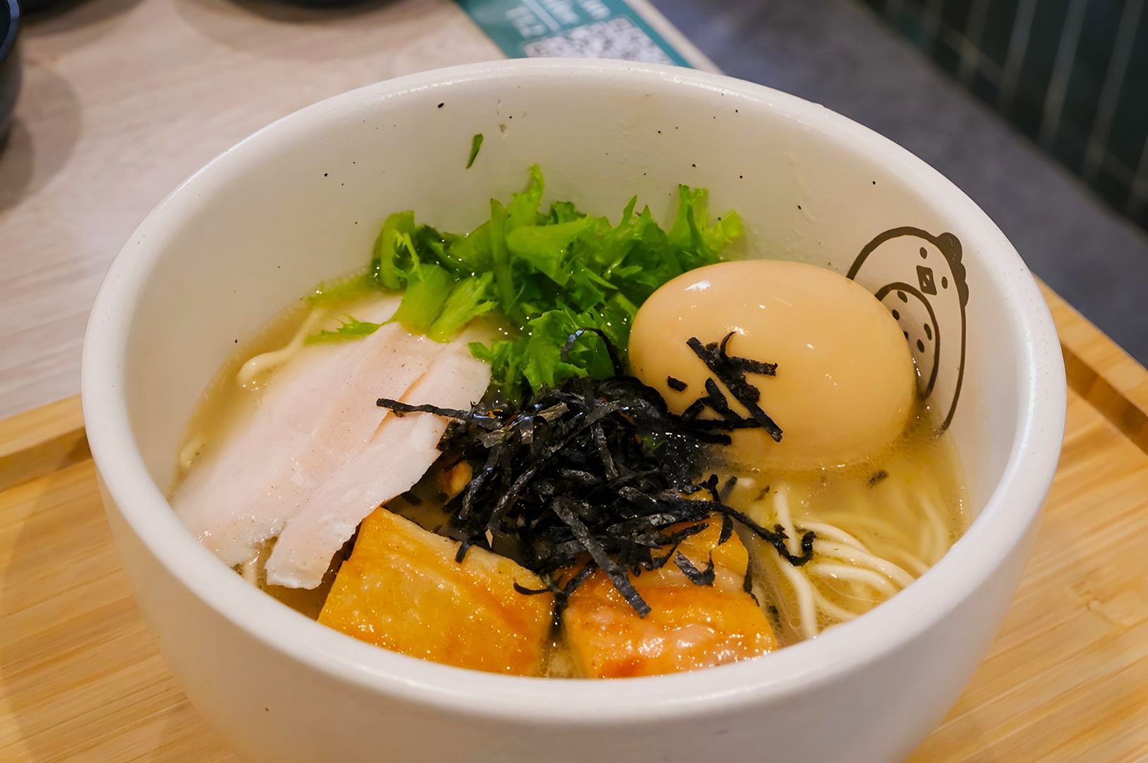 A bowl of ramen with noodles, slices of meat, green lettuce, a soft-boiled egg, black seaweed, and salmon cubes. The dish looks fresh and appetizing.