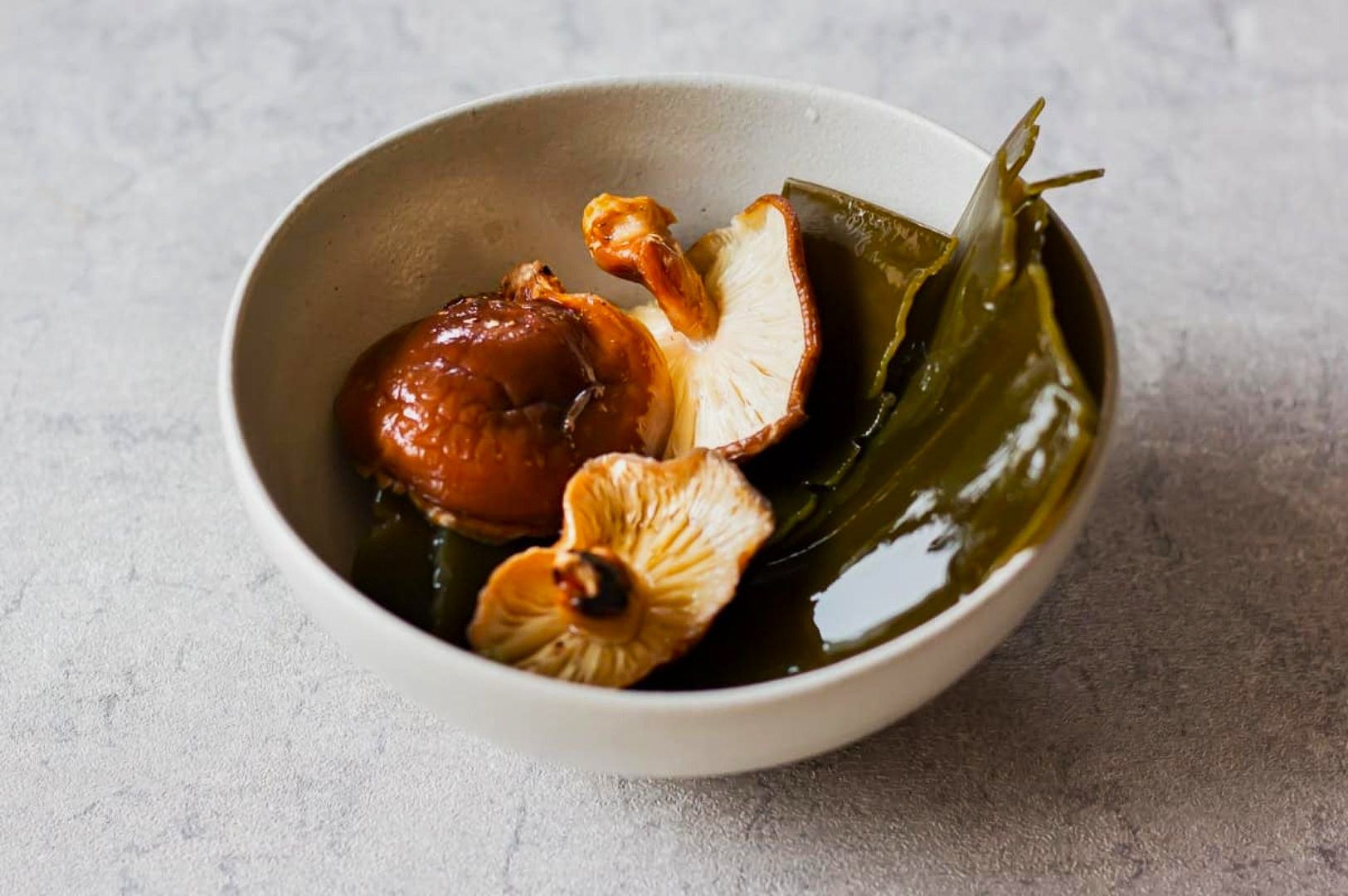 A white bowl on a textured surface holds dark brown mushrooms and green seaweed. The minimalist arrangement conveys a simple, natural aesthetic.