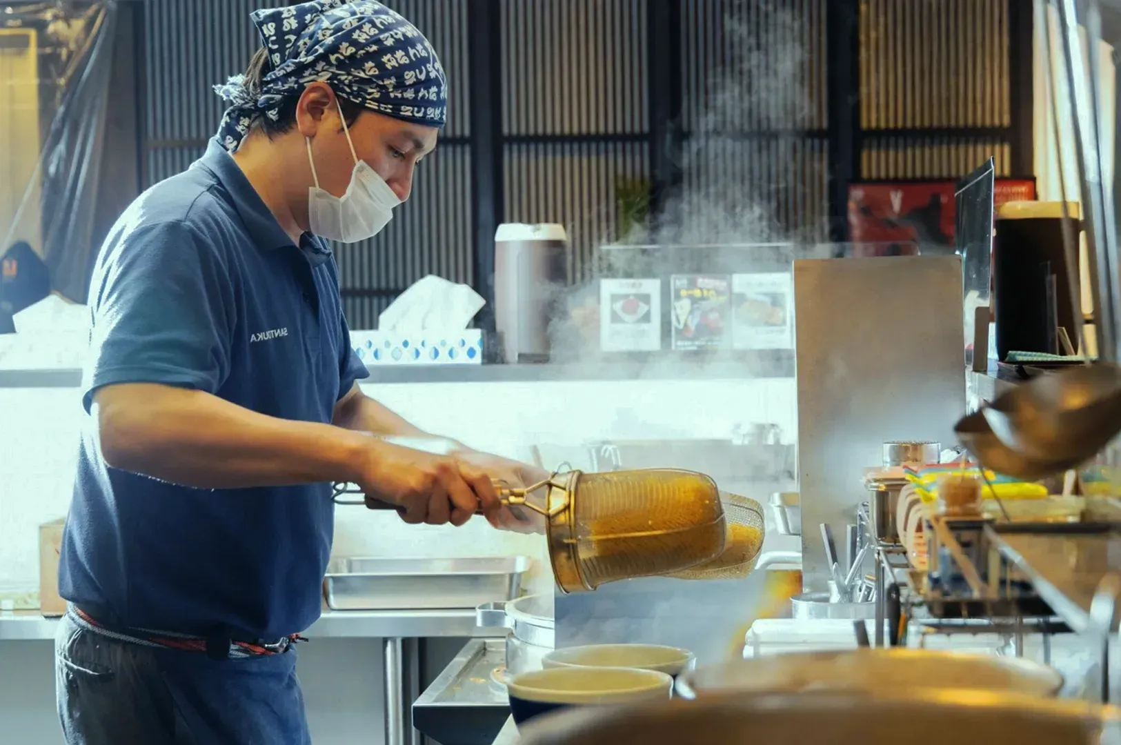 A chef in a blue shirt and bandana, wearing a mask, cooks noodles in a steaming kitchen. The scene conveys focus and dedication in a busy restaurant.