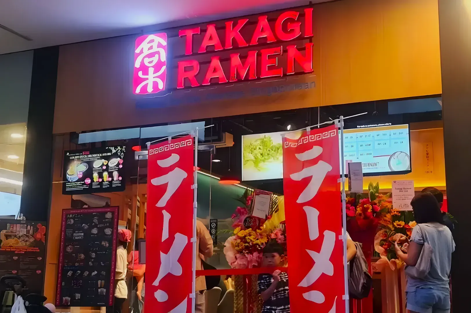 Exterior of Takagi Ramen restaurant with bright red signage. Two red banners with white Japanese text flank the entrance. People and flower stands visible.