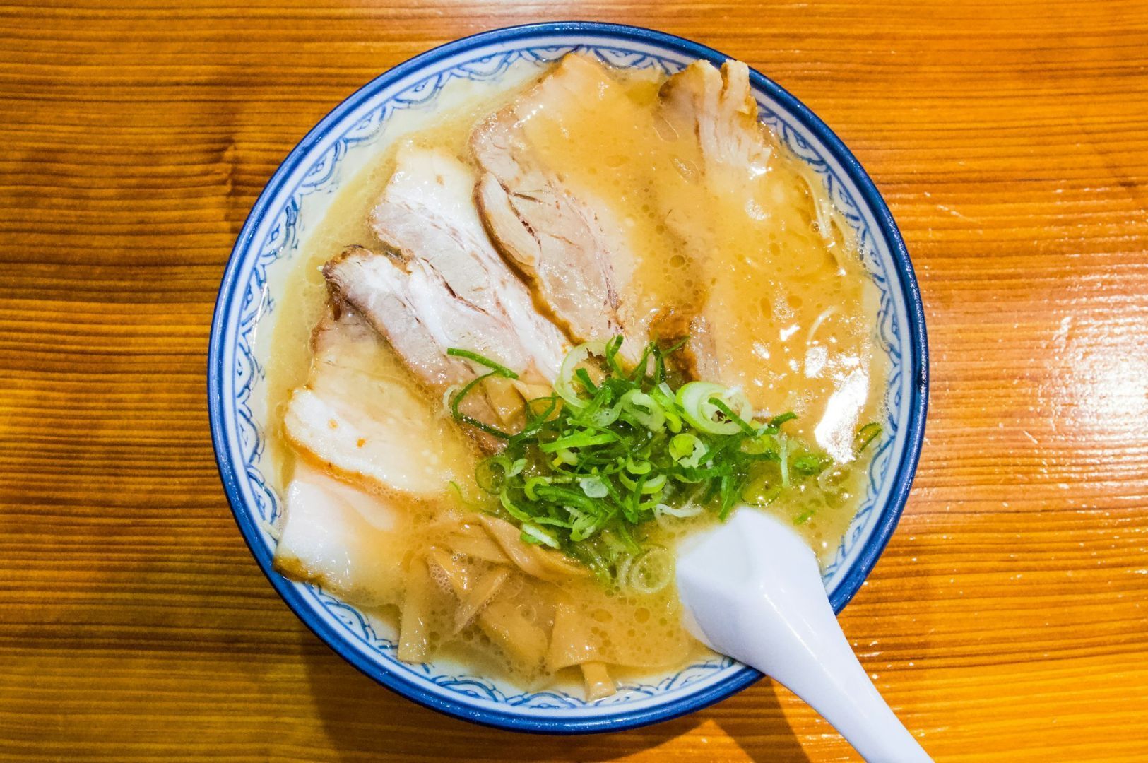 Top-down view of an authentic bowl of Japanese ramen featuring tender chashu pork slices, bamboo shoots, and fresh green scallions in a rich tonkotsu broth, served on a wooden table.