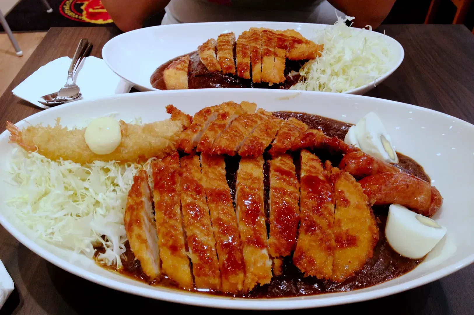 Two plates of Japanese curry feature crispy tonkatsu, shrimp, sliced boiled egg, and shredded cabbage, creating a hearty, savory meal.