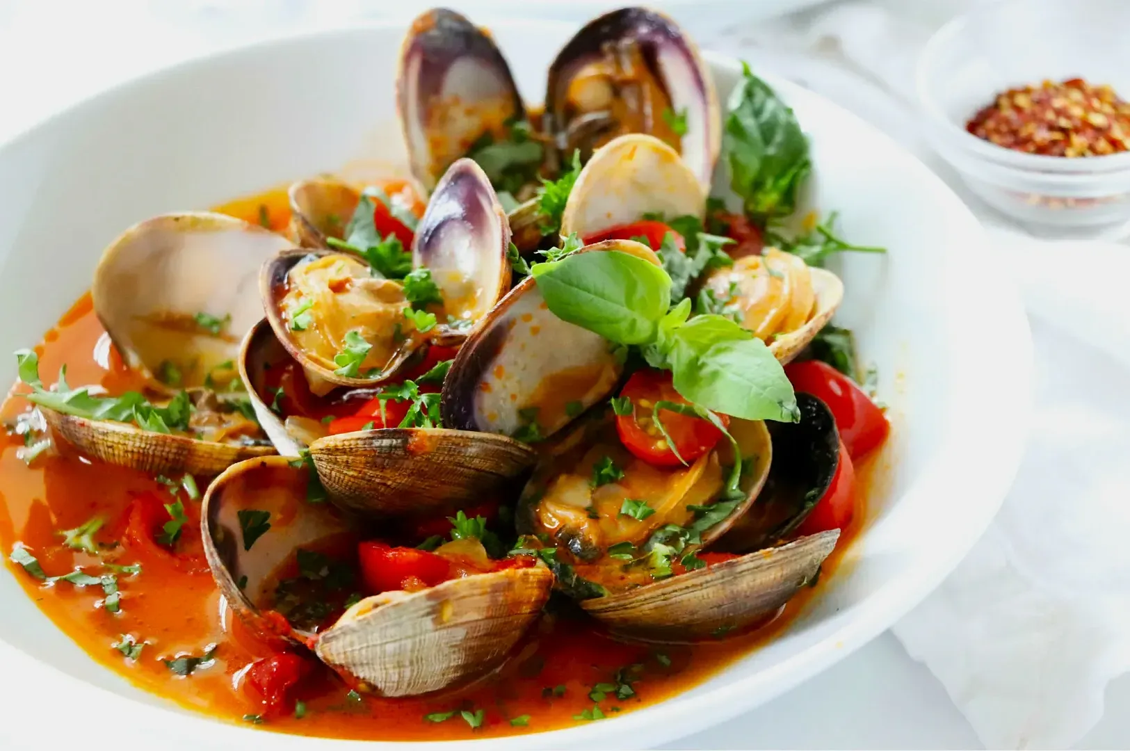 A vibrant bowl of clams in a rich red broth, garnished with fresh basil, chopped herbs, and cherry tomatoes, creating a fresh and appetizing dish.